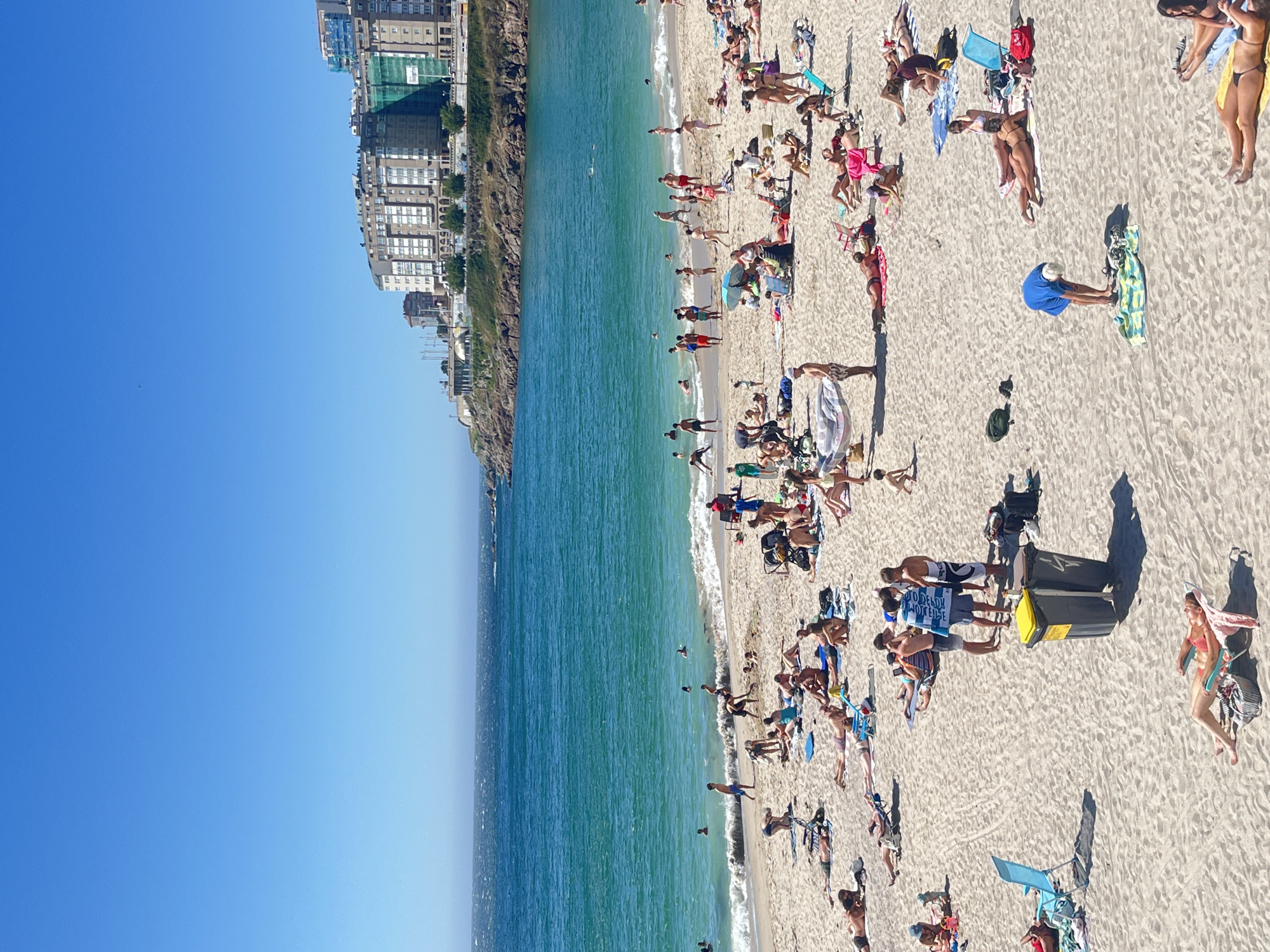 This is a high-angle, wide shot of a crowded beach on a sunny day. The scene is dominated by a wide expanse of sand stretching to meet a vibrant turquoise ocean. In the background, a line of tall, modern buildings runs along the coastline, suggesting an urban setting. The sky is a clear, brilliant blue, indicating fair weather.
Numerous people are scattered across the sand, engaged in various beach activities. Many are sunbathing on towels or lounge chairs, some wearing swimwear, others partially clothed. A few individuals are wading or swimming in the shallow waters near the shore, while others are walking along the beach. The overall atmosphere appears relaxed and leisurely, typical of a summer beach day.
Notable details include the presence of beach umbrellas and towels providing shade. Two large, dark trash bins with yellow lids are situated on the sand, suggesting public amenities. The shadows cast by the people and objects indicate that the sun is high in the sky, likely mid-day. One person is holding a sign with visible text in Spanish, which appears to be "NO DEJEIS MUJERES" - a phrase that translates to "Don't leave women." The location is identified as A Coruña, Spain, which aligns with the urban coastal setting.