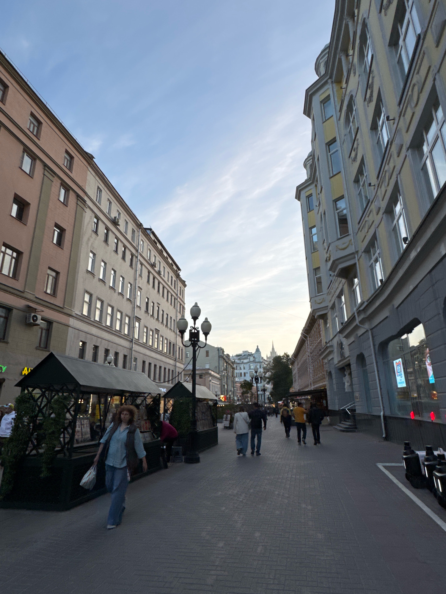 This media file depicts a street scene in Moscow, Russia, likely in the late afternoon or early evening given the soft light and dusky sky. The perspective is low and wide-angled, emphasizing the tall, multi-story buildings lining the cobblestone street. On the left, a pale pink building with numerous windows and air conditioning units stands next to a lighter beige building. On the right, a grander, yellow and gray building with ornate architectural details and arched windows dominates the view.
Several people are walking along the pedestrianized street. In the foreground, a woman with curly brown hair, wearing a blue shirt, vest, and pants, walks towards the camera carrying a bag. Further down the street, other individuals and small groups are seen walking away from the camera. One of the buildings on the left appears to house shops or stalls, with dark green structures and plants visible.
The sky is a soft blue with wispy clouds, suggesting mild weather. The overall atmosphere is calm and urban, with the architectural styles hinting at a European city. No specific text is clearly legible, though some faint signage can be seen on the buildings. The image captures a moment of everyday life on a city street, with people going about their business as the day draws to a close.