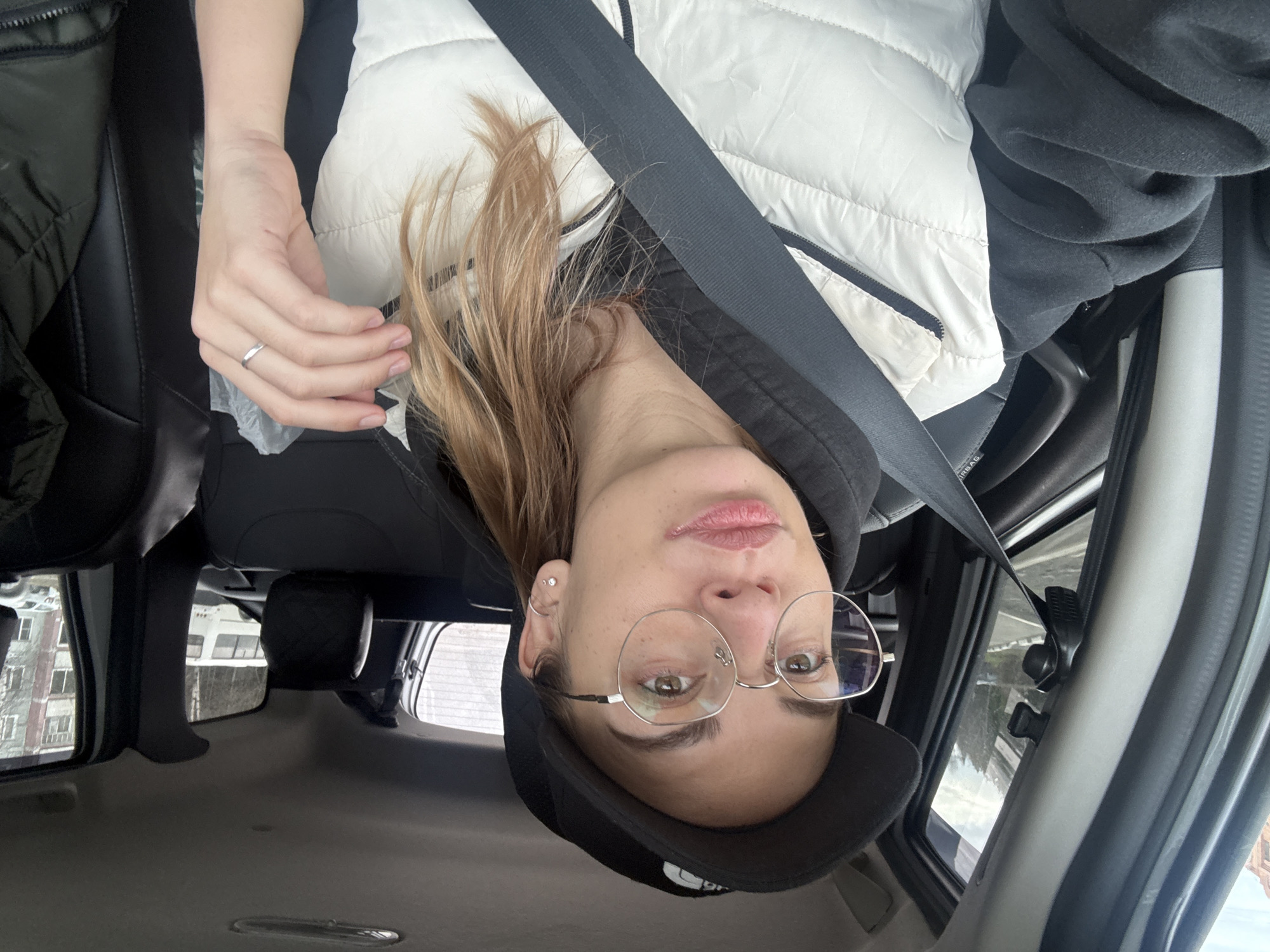 A woman is taking a selfie inside a car. She is wearing a white puffer vest over a black hooded sweatshirt, a black baseball cap, and round eyeglasses. She has blonde hair and is wearing a seatbelt. Her hand is visible with a ring on her finger. The interior of the car is visible, including the seats, headrest, and roof. Through the car windows, a building with many windows and trees can be seen, likely situating the car in an urban area. Given the prompt, the location is in Ust-Ilimsk, Russia.