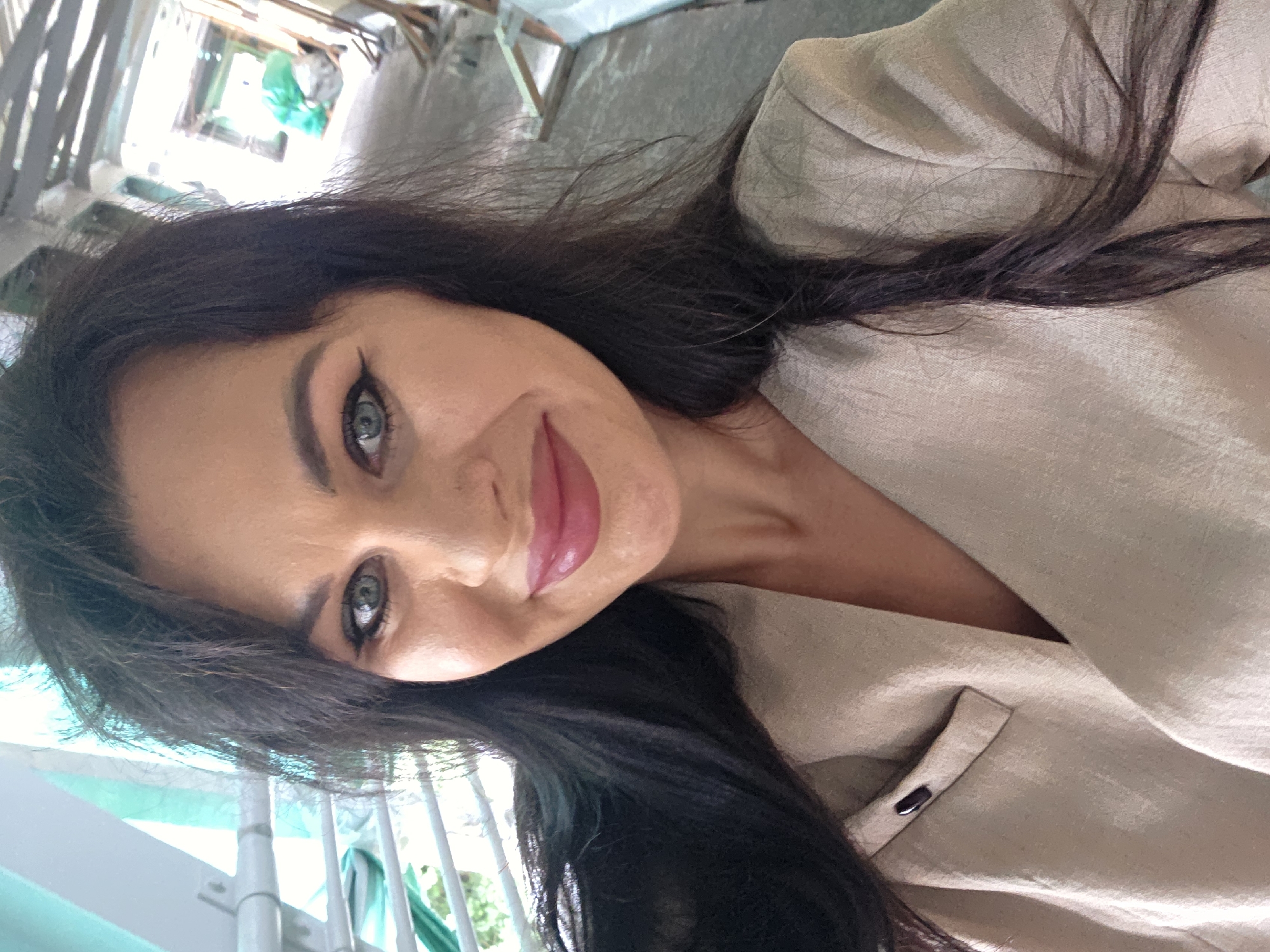 A woman with long, dark hair and blue eyes smiles at the camera. She is wearing a light-colored blazer. The background appears to be an indoor construction site, possibly an overpass or walkway, with metal beams and scaffolding visible. Green tarps are also present in the background, along with some construction materials. The lighting is bright and appears to be natural light coming from overhead.