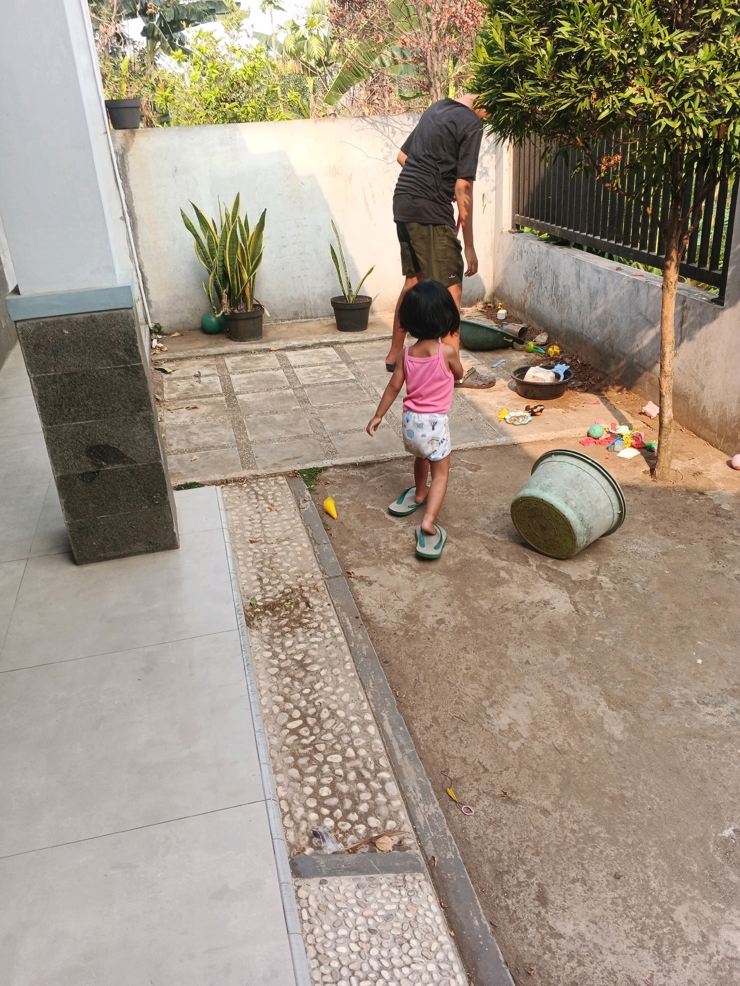 A young girl in a pink tank top and patterned shorts walks away from the camera in a backyard. She is wearing flip-flops. A man in a black t-shirt and olive green shorts stands near a tree and a fence in the background. The ground is concrete and dirt. There are several potted plants, a large overturned pot, a bucket, and various toys scattered around. The environment appears to be a residential area in Kadu, Indonesia.