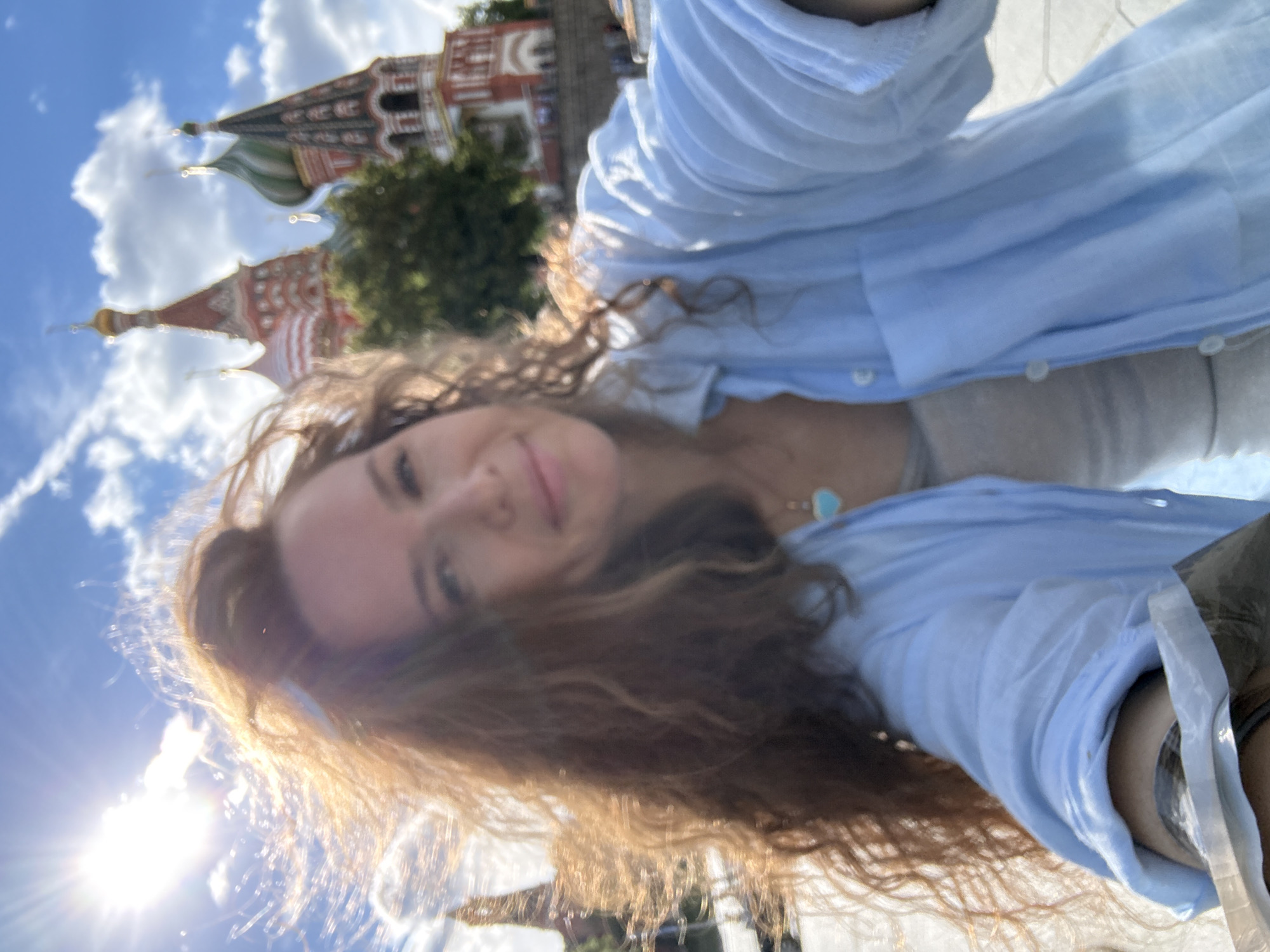 This is a selfie taken by a woman with long, curly brown hair. She is wearing a light blue open button-up shirt over a grey top. The woman is smiling gently and looking towards the camera. In the background, there are distinctive onion-domed buildings, characteristic of St. Basil's Cathedral in Moscow, Russia. The sky is a bright blue with fluffy white clouds. The sun is shining brightly, creating a lens flare effect and casting a warm glow on the woman's hair. The overall scene suggests a pleasant day outdoors in Moscow. There is no visible text in the image.