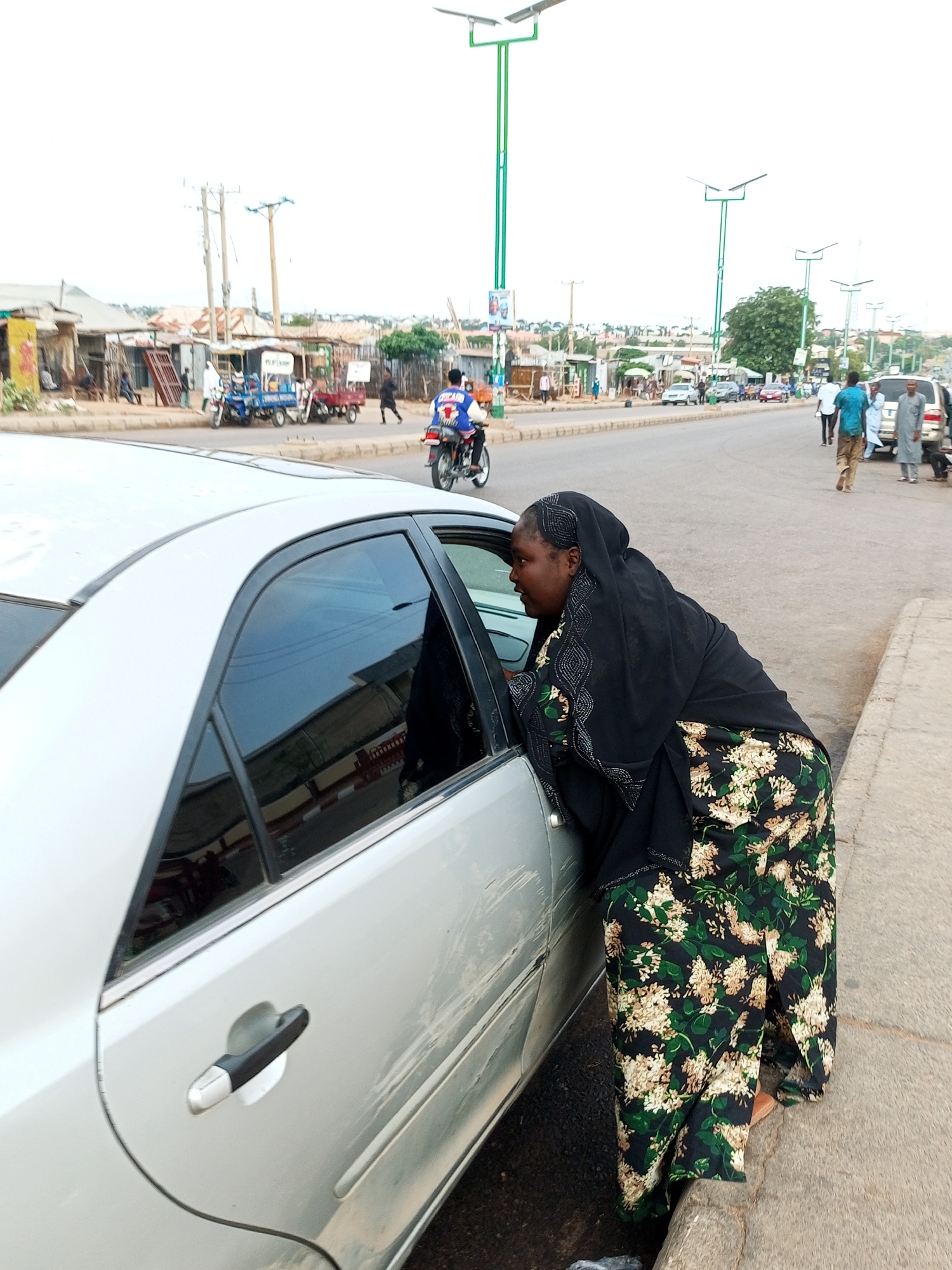 A full-shot, eye-level, medium-light outdoor image shows a woman in a black hijab and a floral skirt leaning into the driver's side of a silver sedan parked on the side of a street in Tumfure, Nigeria. The woman's face is partially obscured by her hijab as she looks into the car. The car's windows are tinted, reflecting the surrounding environment. The street is paved with asphalt and has a concrete curb. In the background, there are several other vehicles, including a motorcycle with a rider, a white SUV, and various smaller vehicles and tricycles. Buildings line the street, with shops and stalls visible. Power lines and streetlights are also present. The sky is overcast.