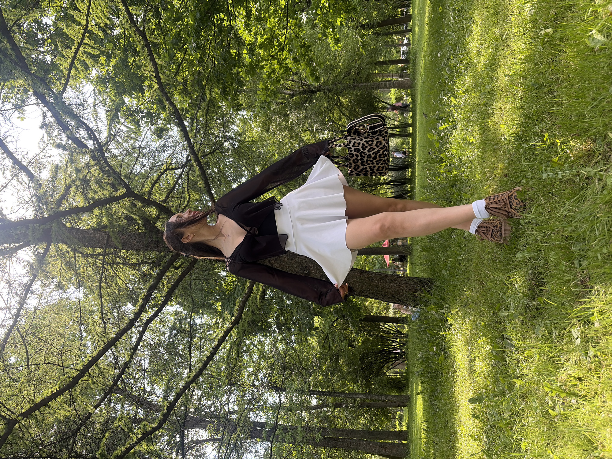 This vertical full shot captures a person standing amidst lush greenery, likely in a park or forest in Saint Petersburg, Russia. The person, viewed from the chest down, wears a black long-sleeved top with a v-neck and a short white pleated skirt. They accessorize with a leopard-print handbag held in their left hand and leopard-print chunky-soled shoes with white socks. Their head is tilted slightly upwards, looking towards the sky, and an earring is visible on their right ear.
The background is dominated by numerous trees with verdant leaves, suggesting a dense wooded area. Sunlight filters through the tree canopy, creating bright spots and shadows on the grass. The ground is covered in green grass with patches of sunlit areas, indicating it is daytime, possibly morning or late afternoon given the quality of light. In the distance, other faint figures can be seen, hinting at other people present in the park. The overall mood appears peaceful and natural. There is no visible text in the image.