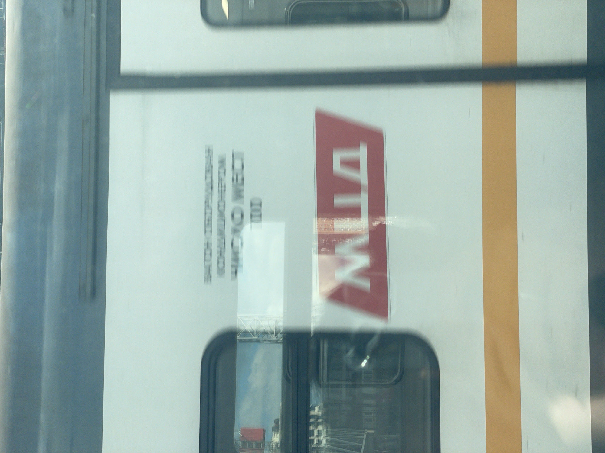 The image shows the side of a white train car in Moscow, Russia. The train has a horizontal orange stripe running across its lower half. There are windows on the train car, and through them, reflections of the surrounding environment, including buildings and the sky, are visible. On the side of the train car, there is a red rectangular sign with a white stylized letter "M" on it, which is the iconic symbol for the Moscow Metro. Above this sign, there is some blurred text in Russian. The image appears to have been taken from another moving vehicle, resulting in motion blur.
