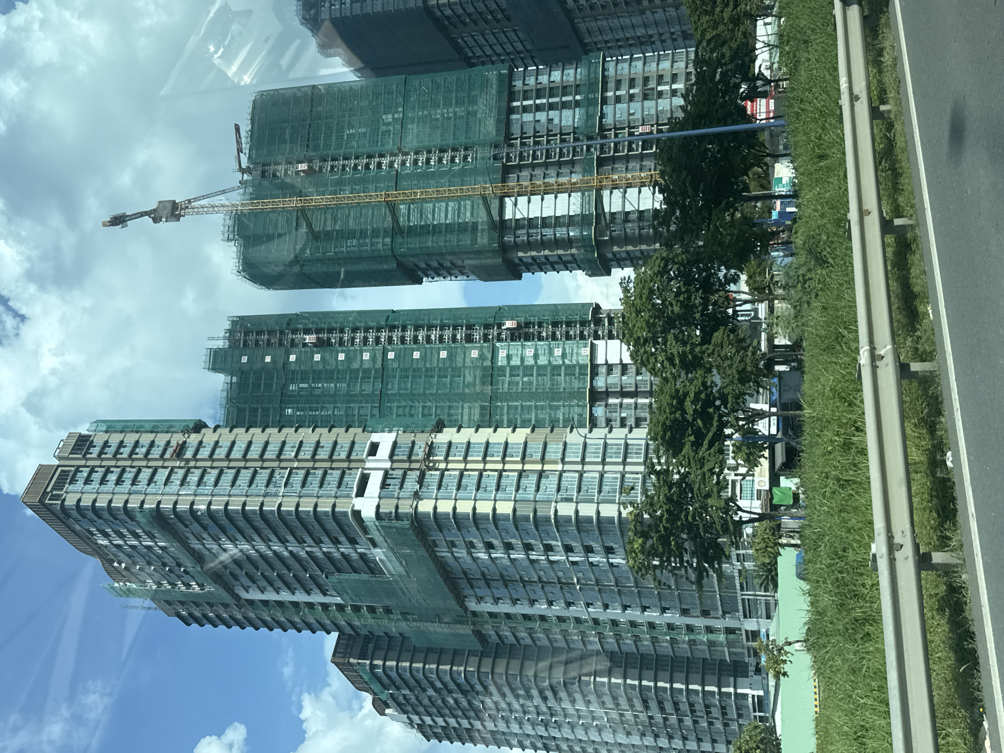 The image shows a partially constructed high-rise building complex in Thủ Đức City, Vietnam. The buildings are tall and modern, with visible glass windows and concrete structures. Several buildings are covered in green netting, indicating ongoing construction. A yellow construction crane is prominently visible, towering over one of the buildings. Trees and greenery are present in the foreground, along with a road and a guardrail. The sky is partly cloudy. The scene appears to be a busy urban area undergoing development.