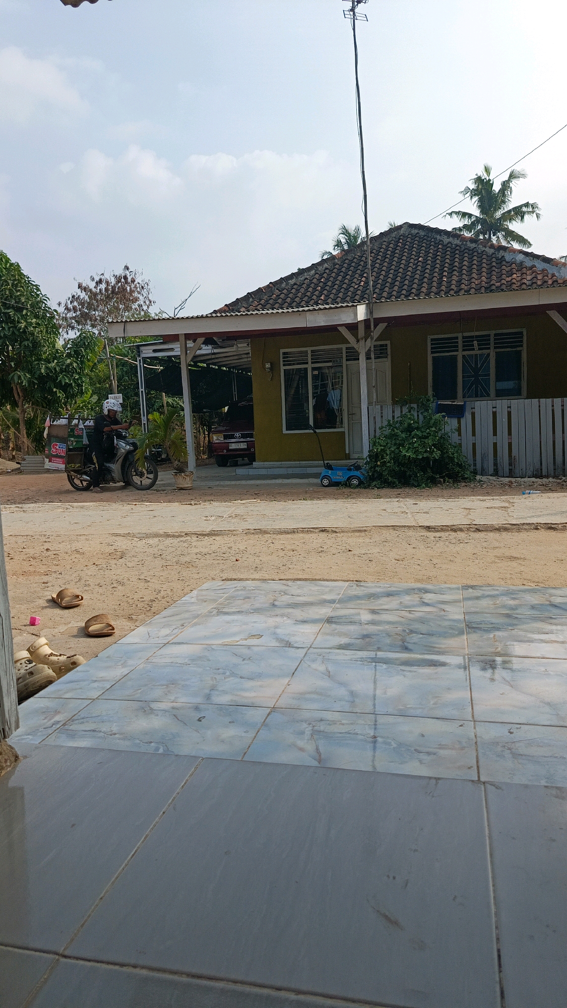 A street scene in the city of Metro, Indonesia, depicts a residential house with a yellow facade and a tiled roof, shaded by a covered porch. In front of the house, a dirt road is visible, with a motorcycle parked on it, carrying a person wearing a helmet. Further down the road, a red car is parked under the porch. Nearby, a blue toy car sits on the ground. The foreground of the image shows a tiled patio area. The sky is partly cloudy.