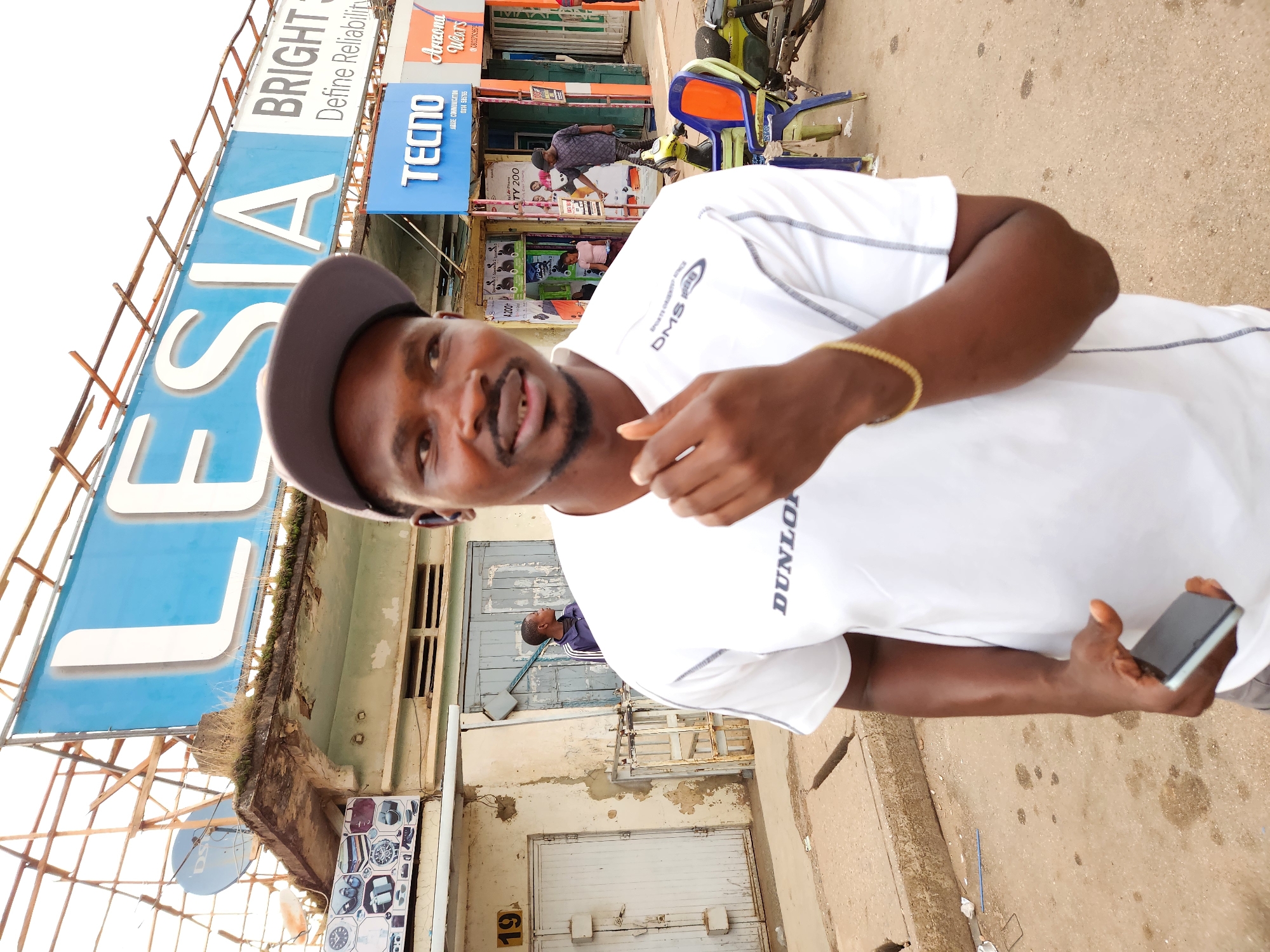A dark-skinned man wearing a white t-shirt with "DUNLOP" and "DMS" logos and a grey baseball cap is standing outdoors in what appears to be a commercial area. He is holding a black smartphone in his left hand. Behind him, a blue sign with white lettering reads "LESIA" and "BRIGHT S Define Reliability". Below this, there are store fronts with signs for "TECNO" and "Arizona Wears". In the background, other people and a motorcycle can be seen. The environment appears somewhat dilapidated, with exposed wooden structures above the signs and peeling paint on the buildings.