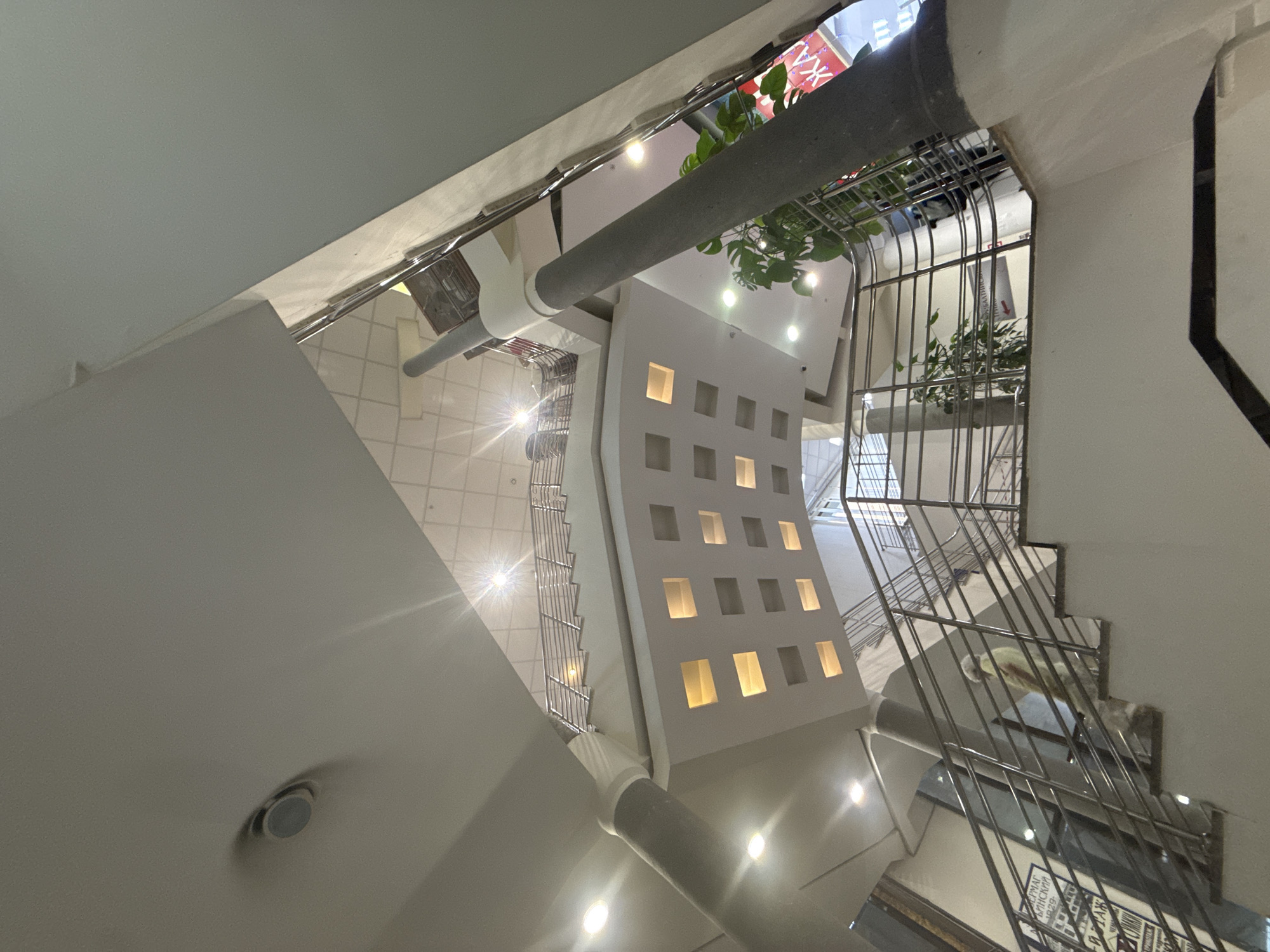 The image depicts the interior of a multi-level structure, potentially a shopping mall or a large public building, in Moscow, Russia. The perspective is from below, looking up towards the ceilings and upper floors.
Architectural elements dominate the scene, including staircases with metal railings, a white structure with square illuminated openings, and cylindrical gray supports or pipes. The color palette is primarily white and gray.
Illumination comes from recessed ceiling lights and the illuminated openings within the white structure. Some plants are visible in the upper portions of the image near the staircases. Faint signage can be observed in the background above the stairs, indicating potential commercial or retail activity. The environment appears clean and well-maintained. There are no people visible.