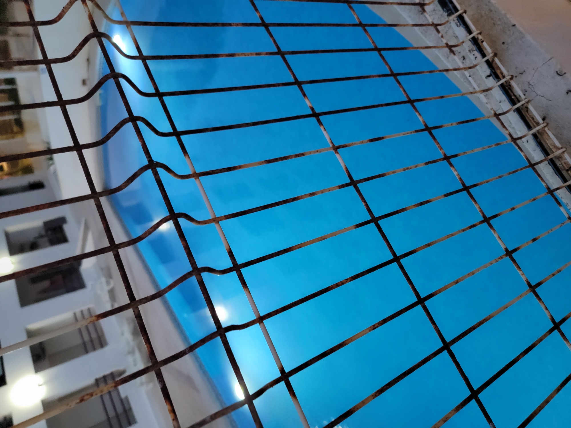 The image captures a view of a bright blue swimming pool at night, as seen through a rusty metal fence. The fence's bars dominate the foreground, creating a grid-like effect. The pool water is illuminated by underwater lights, casting a cool, vibrant glow. In the background, a white building with illuminated windows and balconies is partially visible, suggesting it is a residential or hotel property. The scene is set in the city of Quarteira, Portugal, likely a tourist area given the presence of a swimming pool and the architectural style. There are no people visible in the image.