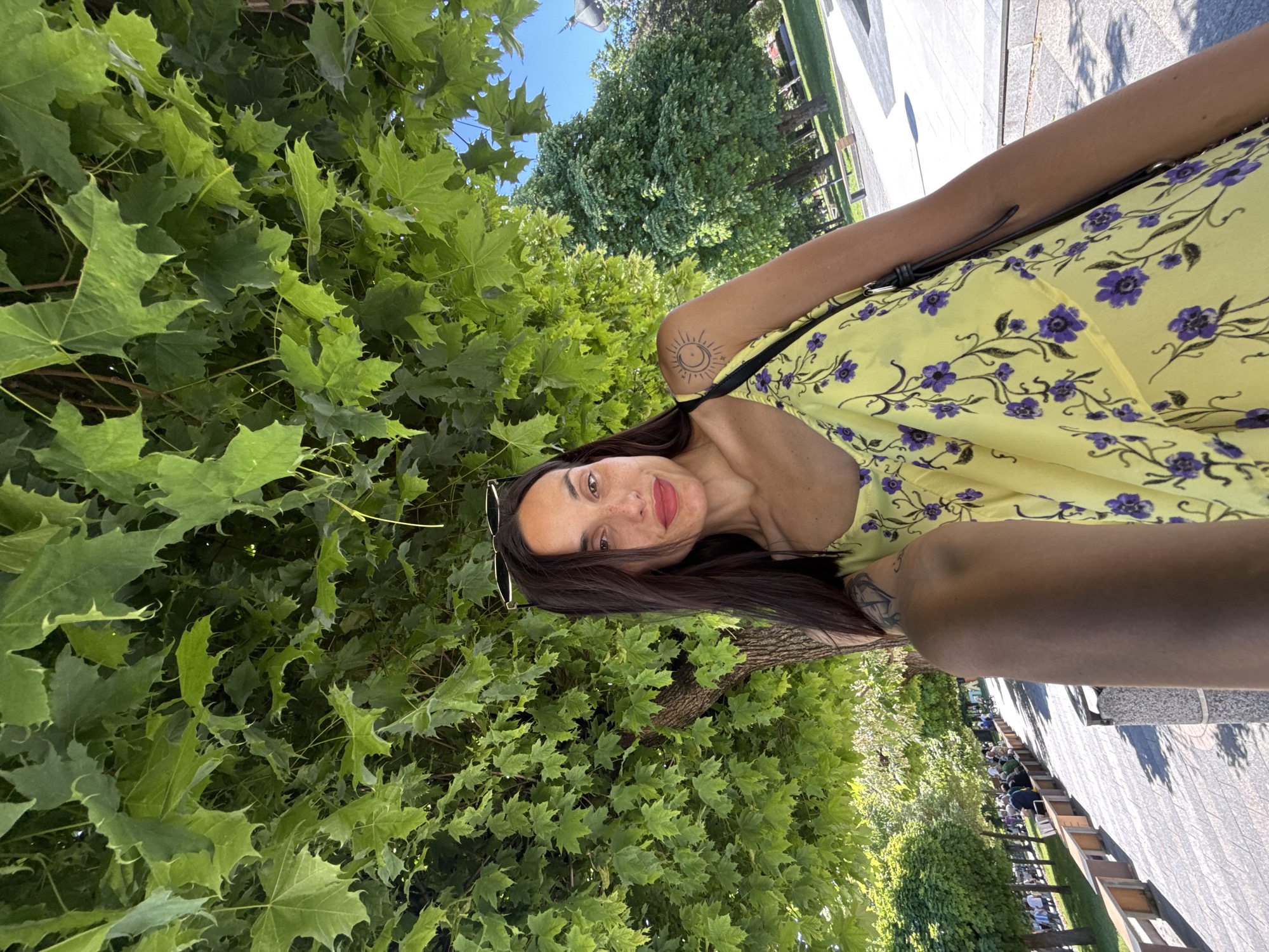The image shows a woman in a yellow floral dress, with sunglasses perched on her head. She stands among dense green foliage, likely a tree. The ground is paved with gray and white stones. In the background, benches line a pathway, and people are seated. The location is in Pleven, Bulgaria.