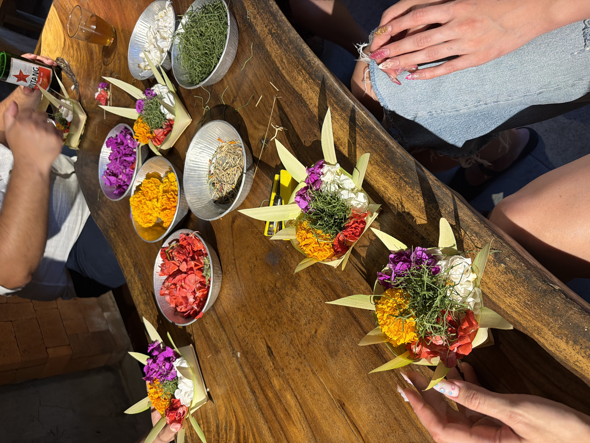 A group of individuals are creating floral offerings on a wooden table in what appears to be an outdoor setting in Mengwi, Indonesia. The table is laden with metal bowls containing various colorful flower petals, green foliage, and dried plant materials. Two people are actively engaged in the craft, one with their hands over a floral arrangement, and another holding a bottle of Bintang beer. Several completed floral offerings, constructed from palm leaves and filled with flowers and greenery, are placed around the table. The individuals are dressed casually, with one person wearing ripped denim jeans and another woman displaying elaborate manicured nails. The lighting suggests it is daytime, with sunlight casting shadows on the table and surrounding brick wall.