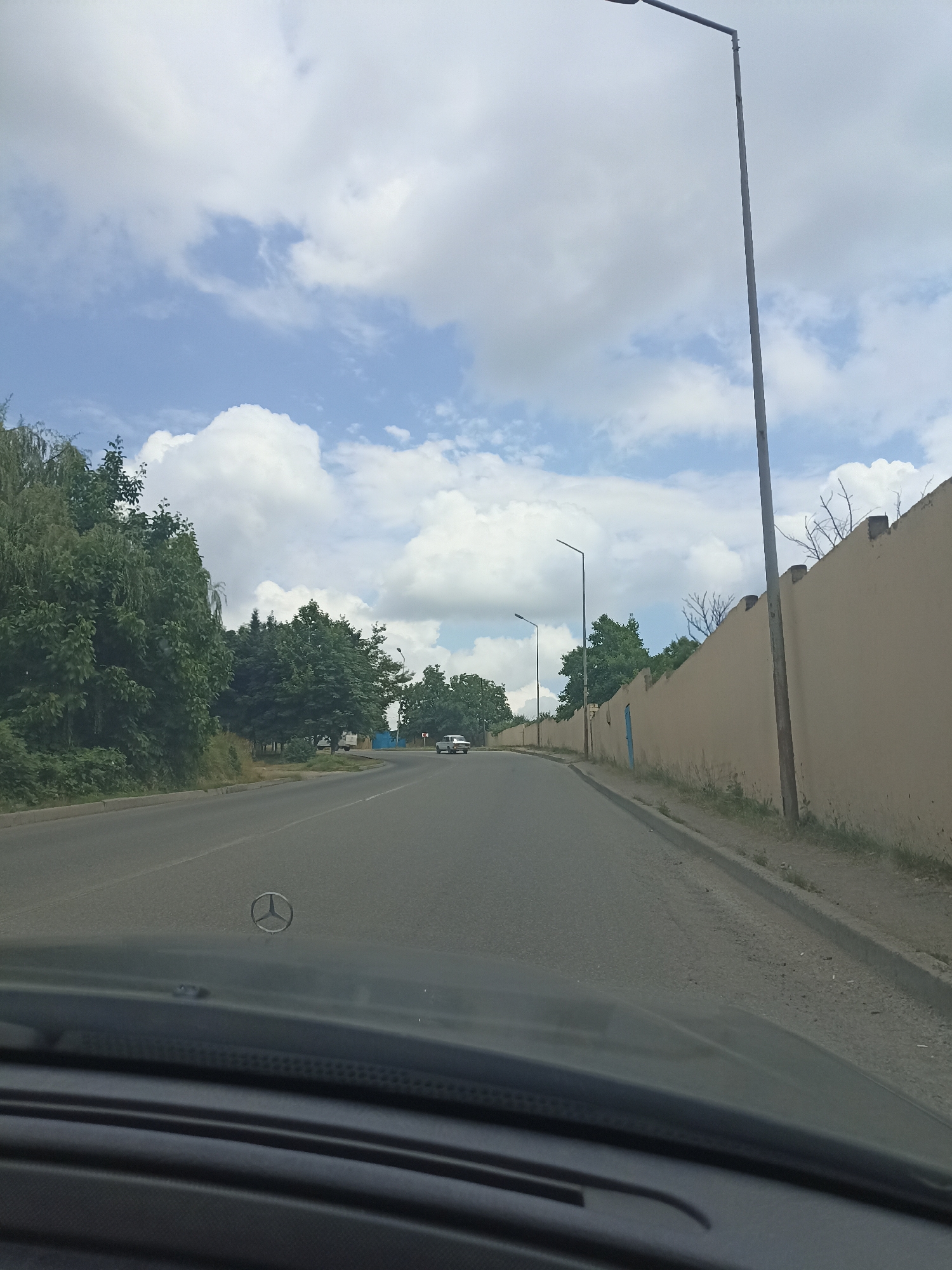 The image captures a view from inside a vehicle, looking out onto a road. The scene appears to be set in Red Sloboda, Azerbaijan. The foreground shows the dashboard and the Mercedes-Benz emblem on the hood of the car. The road ahead curves to the left, with a concrete barrier on the right side and trees and vegetation on the left. A long, tan-colored wall runs along the right side of the road, with a lamppost in front of it. The sky is partly cloudy with large white cumulus clouds against a blue backdrop. Several other lampposts are visible along the road, and a few vehicles are in the distance. The overall impression is that of a drive through a suburban or semi-rural area.