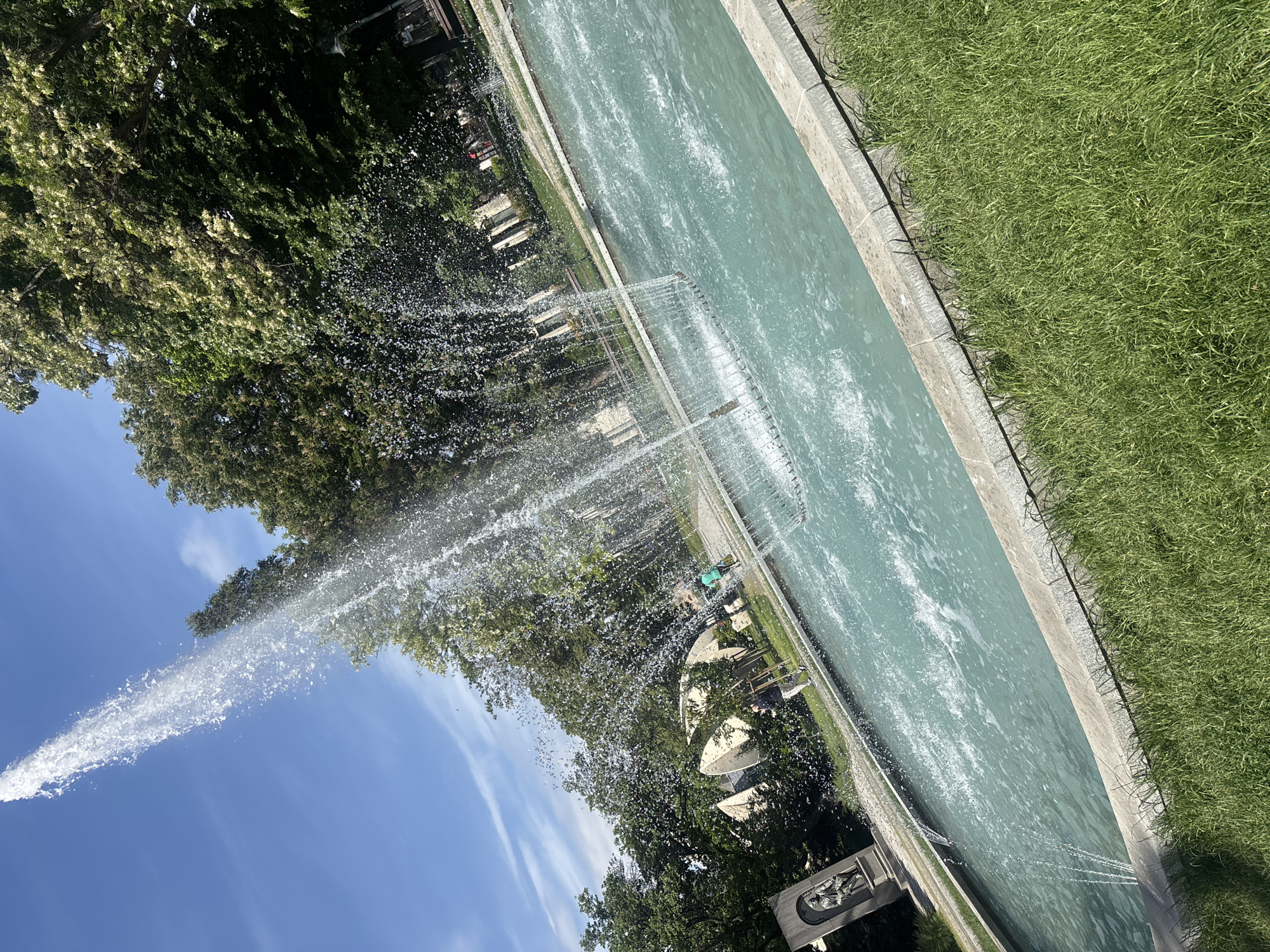 A tilted view of a fountain in the city of Plovdiv, Bulgaria, is seen on a sunny day. The fountain is rectangular, with pale green water and a concrete border. It is situated between lush green grass on the right and trees on the left. A large water jet shoots upward from the fountain, splitting into numerous droplets as it rises. Benches and sunshades are visible along the fountain's edge. In the lower left, a statue sits near the fountain's base.