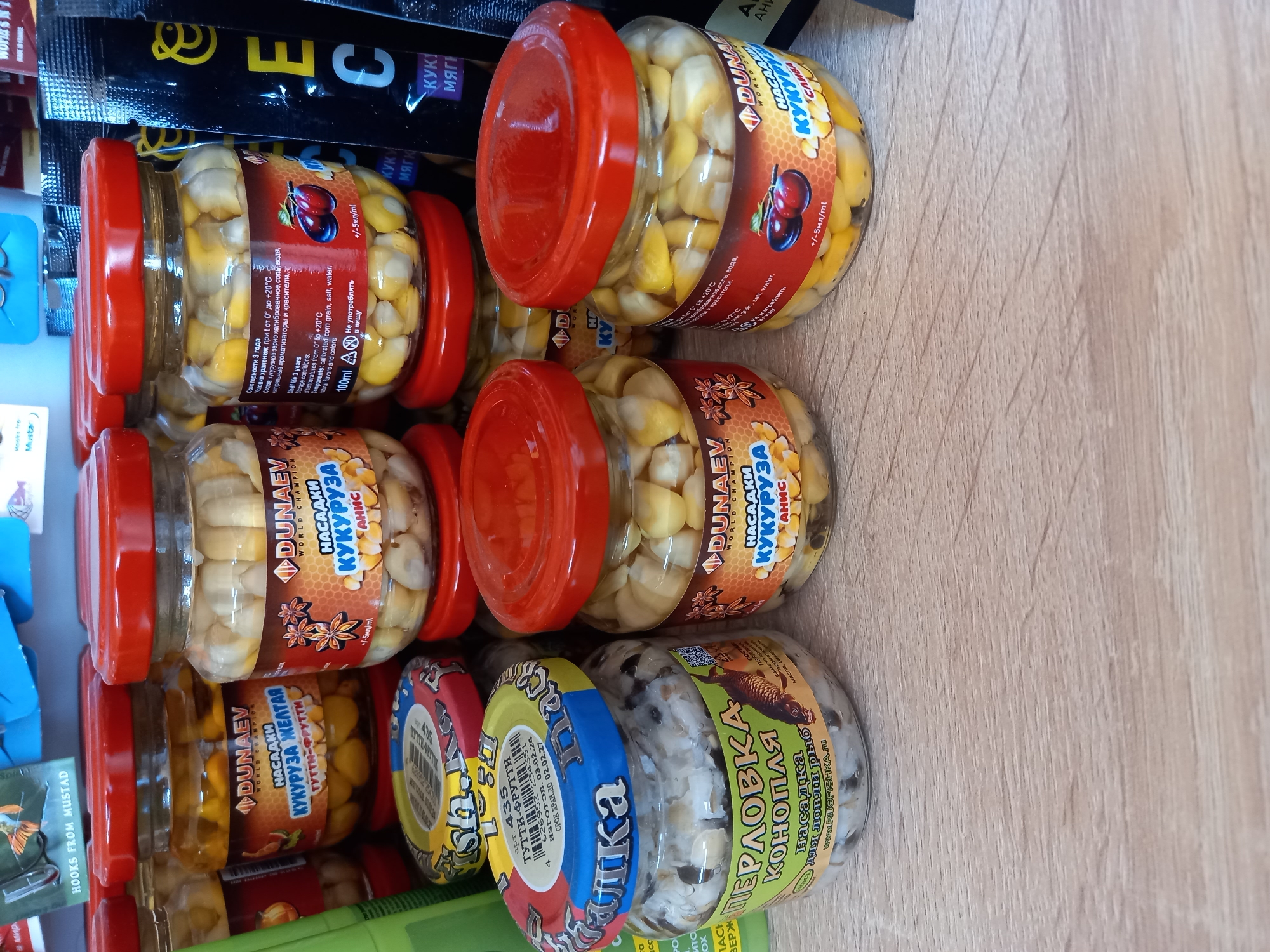 A variety of glass jars, mostly containing bait for fishing, are arranged on a light brown wooden surface. The jars are of different sizes and shapes. The majority of jars have red lids and are filled with yellow corn kernels and clear liquid, labeled "DUNAEV" with different flavors like "ANIS" and "TUTTI-FRUTTI." One jar has a blue and red lid and contains a white substance with dark specks, labeled "ПЕРЛОВКА КОНОПЛЯ" in Russian, indicating pearl barley and hemp. The background is slightly blurred, showing parts of other products, including packaging in black, blue, green, and yellow. The jars are stacked and positioned in a way that labels and contents are visible.
