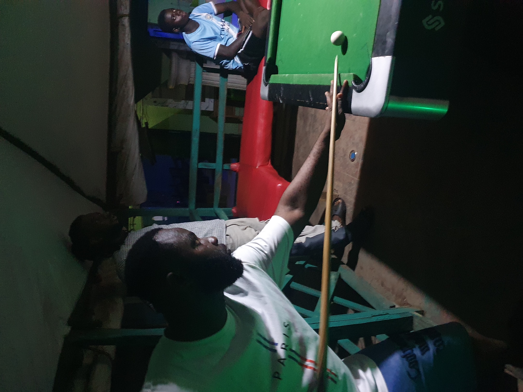 The image depicts a group of individuals engaged in a game of billiards in Lokoja, Nigeria. In the foreground, a man wearing a white t-shirt with "PARIS" printed on it is actively playing, aiming a cue stick towards a white billiard ball on the green felt surface of the pool table. He is positioned at an angle, facing the table. Behind him, another man in a striped shirt stands, observing the game. To the right, a young man in a light blue t-shirt and dark shorts is seated on a red bench, also watching the game. The environment appears to be an outdoor or semi-outdoor setting, possibly a bar or recreational area, with a wooden structure above and green railing visible. The lighting suggests it is evening or night. The pool table itself is a standard size with green felt and visible cushions. The overall scene conveys a casual, social gathering centered around the game of billiards.