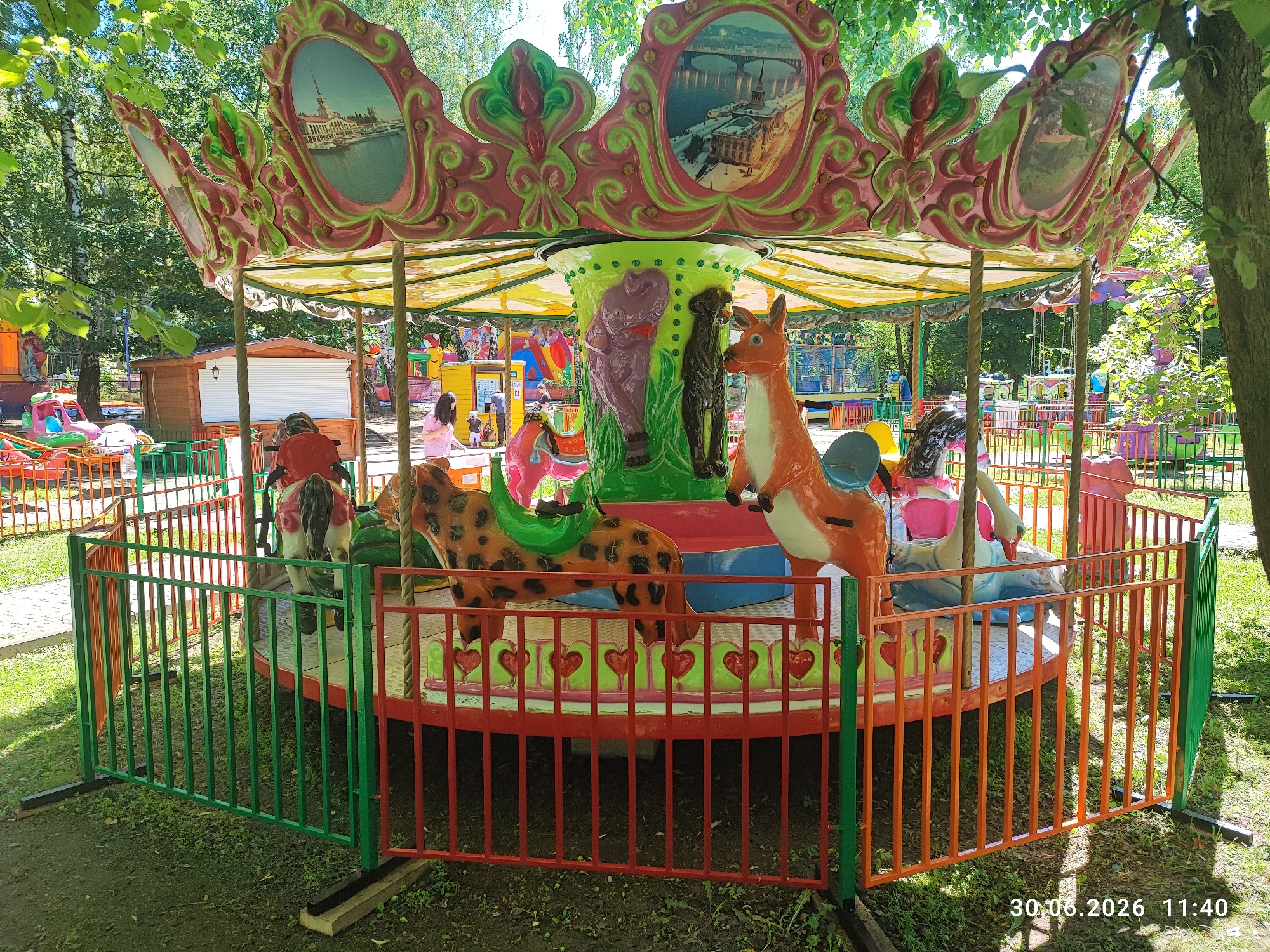 A vibrant carousel is set up outdoors in a park-like area. The carousel features a colorful canopy adorned with decorative trim and oval paintings depicting cityscapes. The central pole of the carousel is decorated with sculpted animals, including a purple hippopotamus and a black dog, alongside an orange kangaroo. The carousel itself has various animal figures for riders, such as a leopard and a swan.
The carousel is surrounded by a colorful metal fence, with sections of green, red, and orange. Beyond the carousel, other amusement park rides are visible in the background, including a smaller carousel and what appears to be a kiddie ride with a pink car. There are also some wooden structures, possibly ticket booths or game stalls.
In the foreground, the ground is covered with grass and some patches of dirt. The lighting suggests it is daytime, with bright sunlight filtering through the leafy trees that surround the area. A few people are visible in the background, seemingly enjoying the park. The overall scene is cheerful and lively, indicative of a fun day at an amusement park.
A timestamp in the lower right corner reads "30.06.2026 11:40". The location context provided is Lytkarino, Russia.