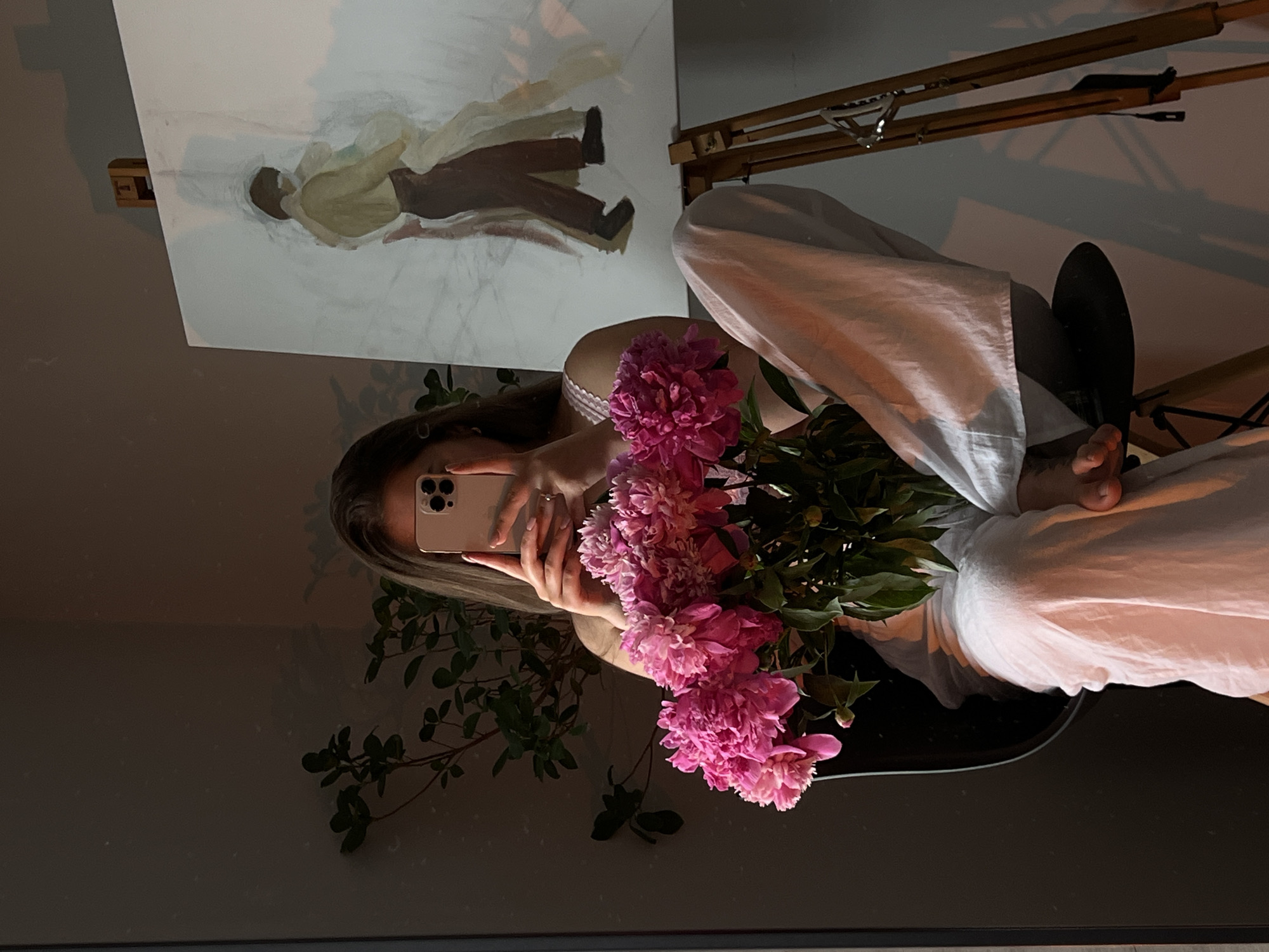A person is sitting on a black chair, holding a pink phone in their hands, taking a selfie. They are wearing a white camisole and loose white pants. In their lap, they are holding a bouquet of pink peonies. To their right, an easel stands with a partially completed painting of two figures. A plant is visible to the left of the person. The setting appears to be an indoor space, possibly an artist's studio or a room with artistic elements. There is no visible indication of the city of Ternopil, Ukraine, in the image.