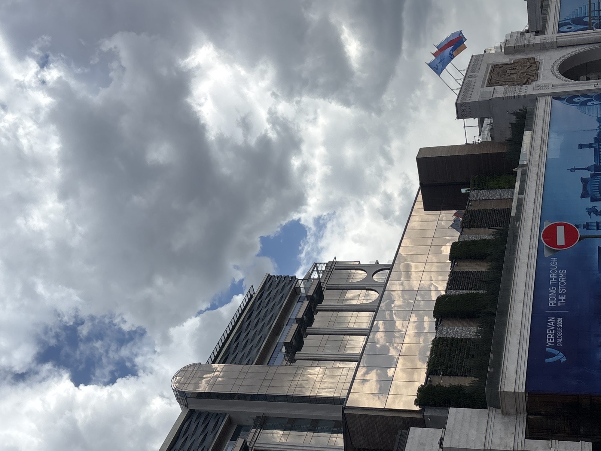 The image shows a view of buildings in Yerevan, Armenia, under a cloudy sky. A modern building with reflective glass and rounded balconies is on the left. A larger building with a banner reading "YEREVAN DIALOGUE 2026 RIDING THROUGH THE STORMS" is partially visible on the right. Russian and Armenian flags are flying on top of the building.