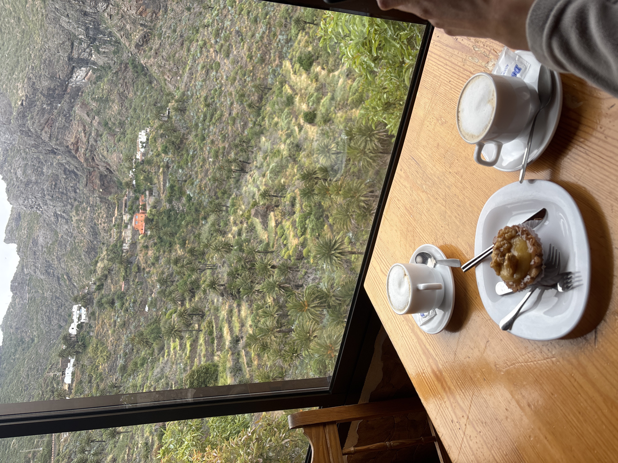 A high-angle interior shot depicts a wooden table next to a window with a scenic view. On the table, there are two white cups of coffee with frothy tops, each resting on a saucer with a spoon. One cup also has a sugar packet next to it. There is also a white plate with a pastry, a knife, and a fork. The window frames a view of a mountainside with sparse vegetation and a few buildings nestled into the landscape. Palm trees dot the scene. Part of a person's arm and sleeve is visible in the upper-right corner. The location appears to be Buenavista del Norte, Spain, based on the mountainous terrain and vegetation.
