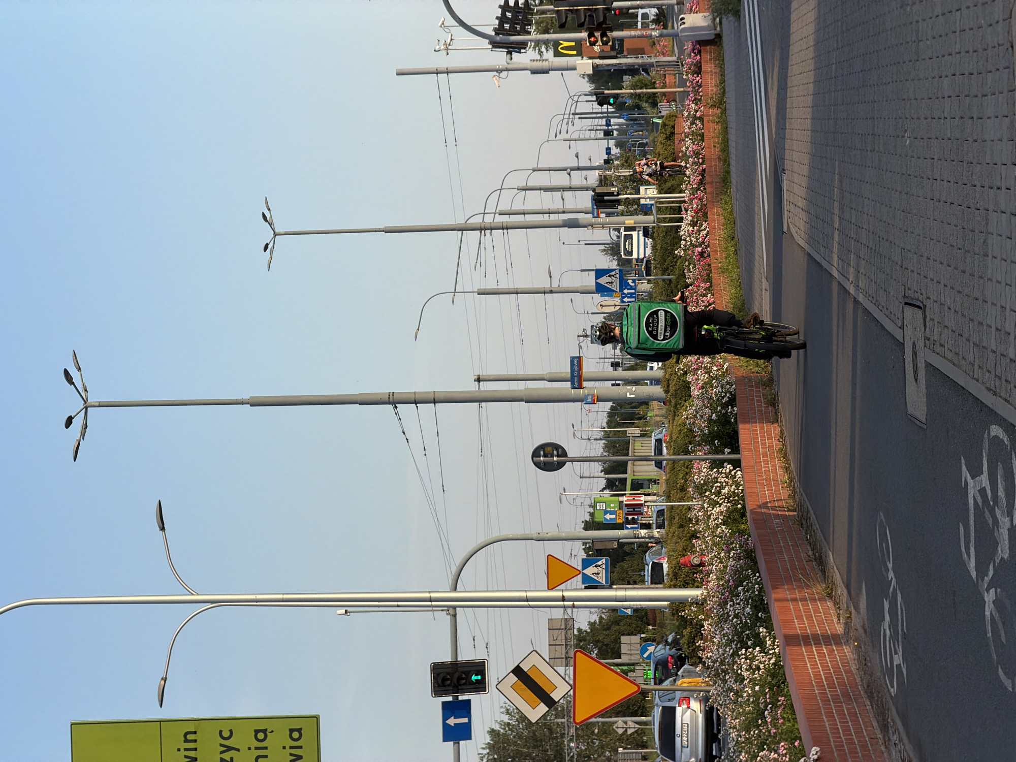 A wide shot captures an urban scene with a cyclist wearing a green "Uber Eats" backpack riding a bicycle on a bike path. The cyclist is moving away from the camera, heading into the distance on a paved path bordered by a low brick wall and a flowerbed. In the background, traffic lights display a green signal, and several street lamps with multiple lights are visible, along with power lines and road signs. Cars are parked and moving on the road. The sky is clear and light blue. The location is an urban environment in Unknown, Unknown.