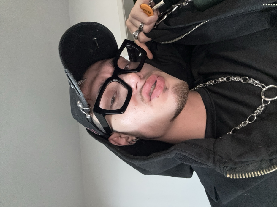 The image displays a close-up, chest-up portrait of a single individual, appearing male, captured indoors against a plain, light grey wall. The individual has light skin, a short goatee beard, and full lips forming a slight pout. They are wearing large, angular black-framed glasses. A black baseball cap adorned with silver-colored metal rings and spikes covers the upper part of their head. The individual is dressed in a black hooded jacket or sweatshirt, partially unzipped, over a black t-shirt. A silver-colored chain necklace with varying link sizes and a prominent O-ring hangs around their neck. The left earlobe features a silver-colored hoop earring. The individual's right hand, which has a silver-colored ring on the ring finger (possibly a coiled snake design), is positioned near the right side of the jacket, holding a keyring with several keys, one featuring an orange cap. The individual is looking directly into the camera. The generic background provides no specific visual cues to identify a location within Sofia, Bulgaria.