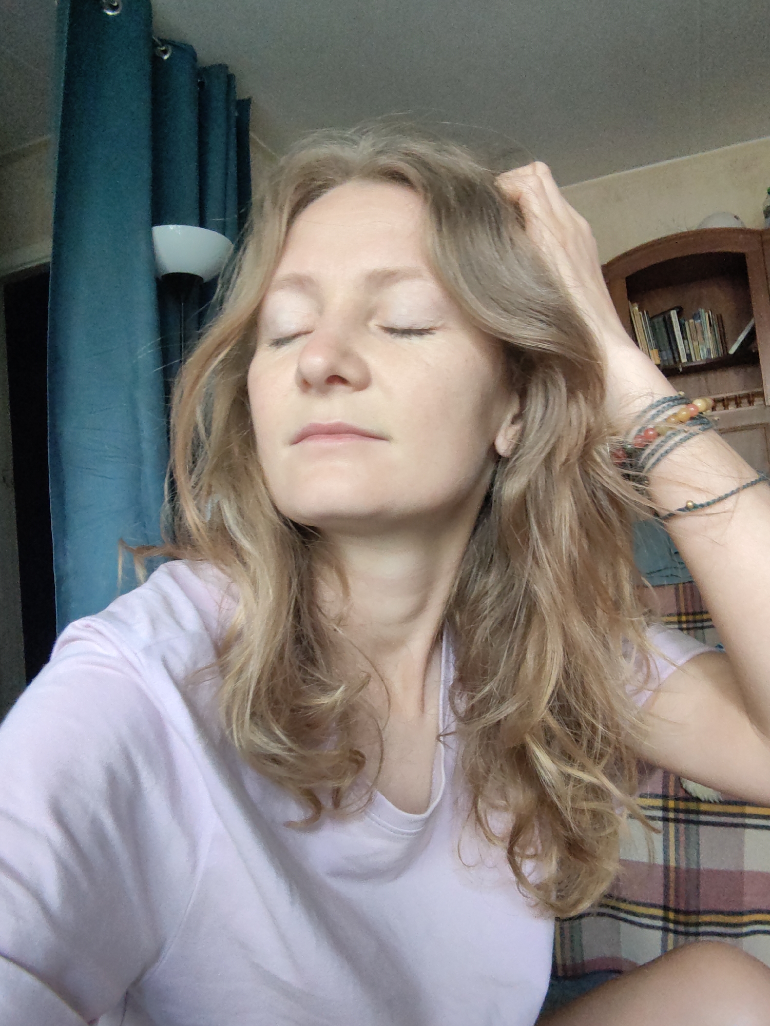 A light-skinned woman with long, wavy blonde hair, wearing a light pink t-shirt, is sitting indoors with her eyes closed. Her right hand is raised, fingers touching her head. She is wearing multiple bracelets on her right wrist. Behind her, on the left, are teal curtains and a lamp. To her right is a wooden bookshelf filled with books. Behind her shoulder is a plaid fabric. The location is not Moscow, Russia.