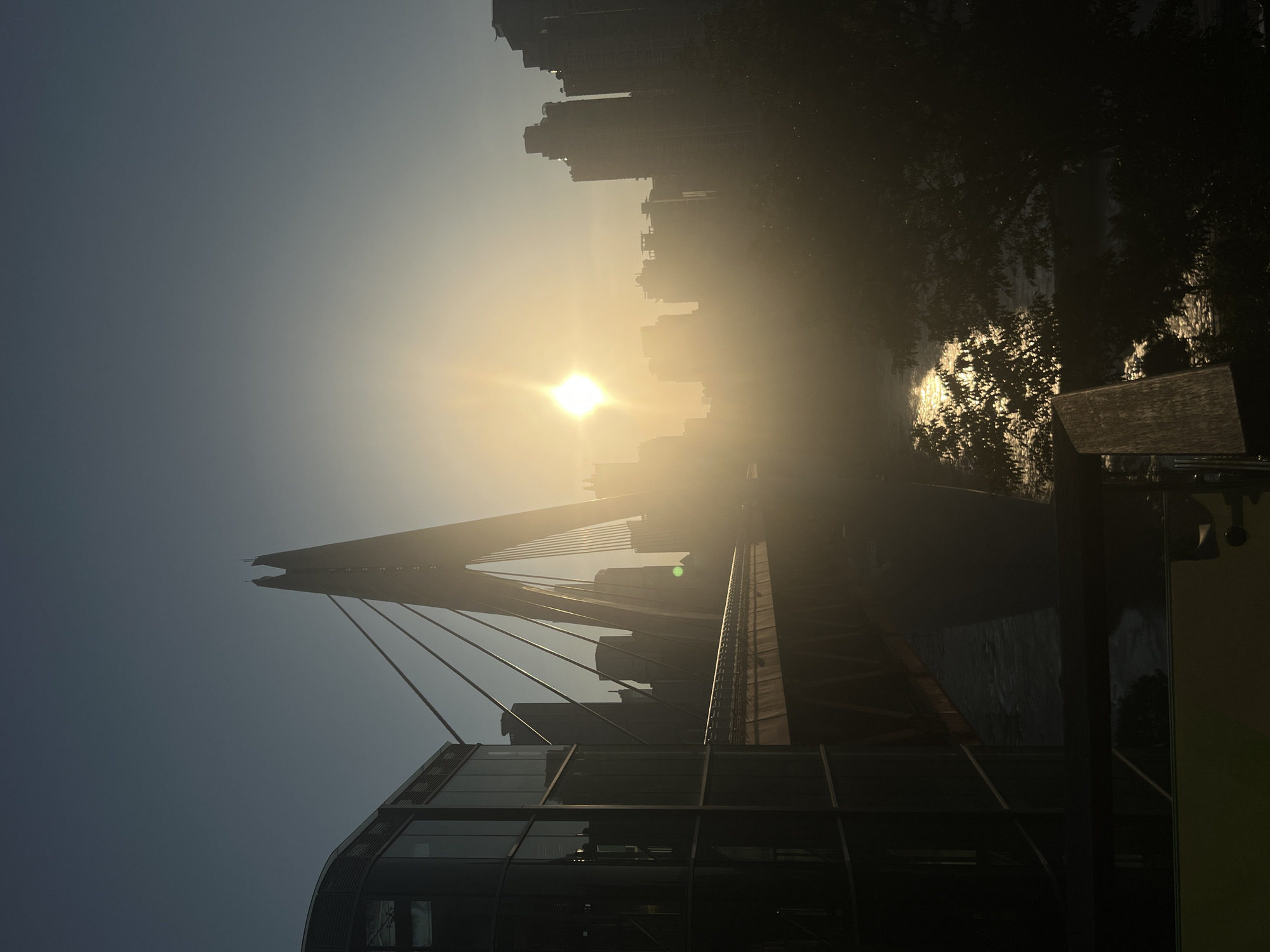 An outdoor, horizontal, medium shot captures a cityscape at sunrise. The sun, a bright, starburst-shaped orb, dominates the center of the frame, casting a warm glow through a hazy atmosphere. In the foreground to the left, a modern glass-fronted building stands tall, its reflective surfaces mirroring the dim sky. Extending from it is a suspension bridge, its steel cables and structure silhouetted against the bright sky. The bridge stretches across a body of water, its surface shimmering with reflected sunlight. Behind the bridge, a cluster of towering city buildings forms a hazy silhouette against the sunrise. The scene is devoid of people and animals. The location is identified as Nan'an District, China.