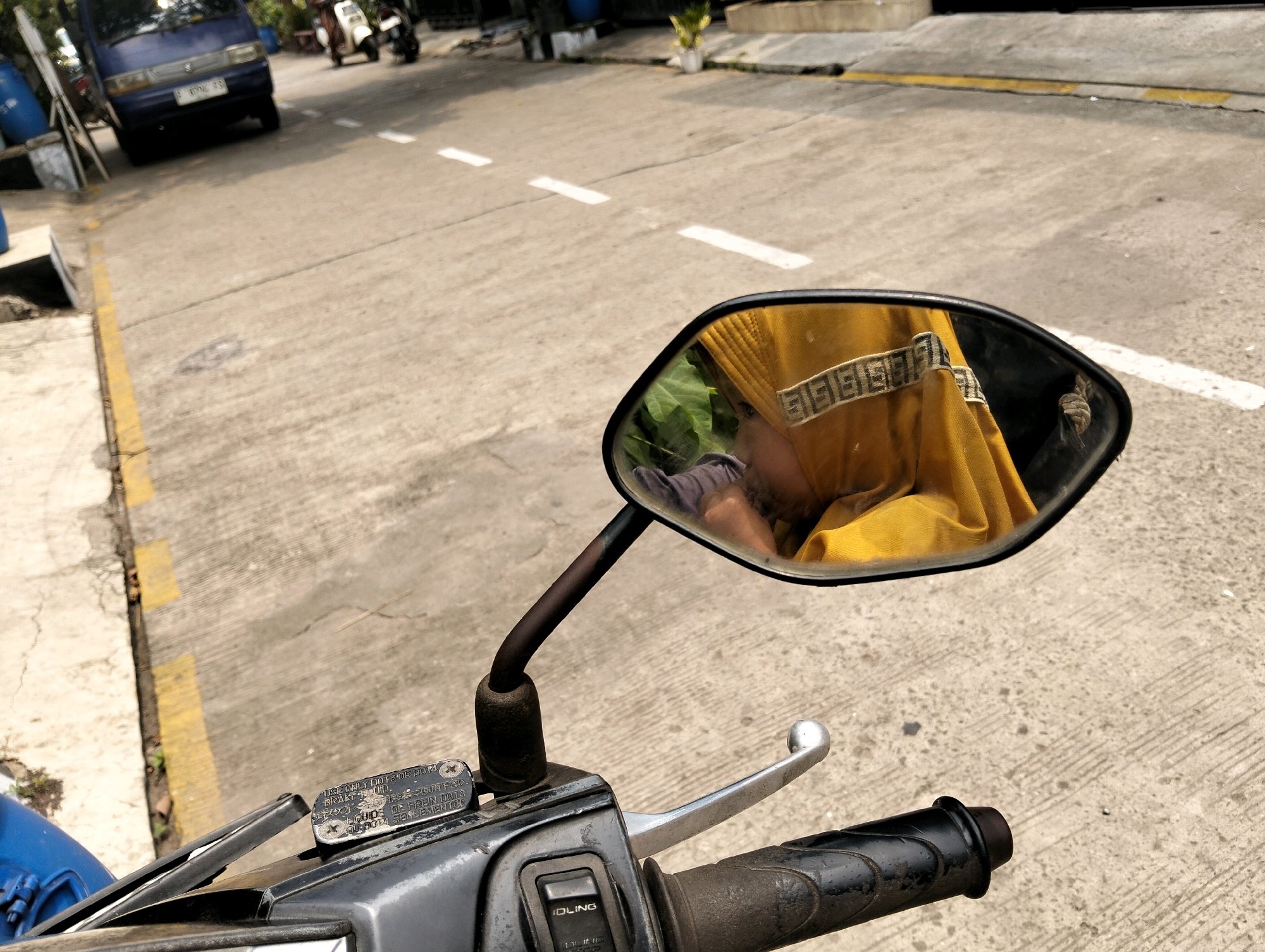 The image is a first-person perspective of someone sitting on a motorcycle in Cileungsi, Indonesia, on a paved road. The road has white dashed lines indicating lanes and a yellow line along the curb. A blue van is visible in the background, along with two scooters parked on the side of the road. A side mirror on the motorcycle reflects the rider, who is wearing a yellow garment. The rider's hand is visible on the handlebar, which includes the brake lever and a switch labeled "IDLING." The environment appears to be outdoors on a sunny day.