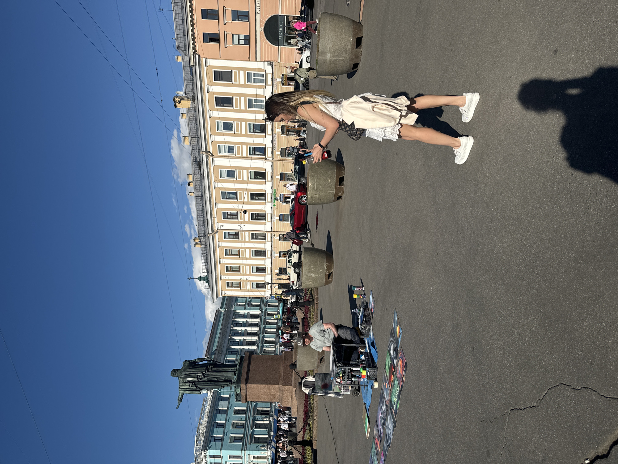The image shows a street scene in Saint Petersburg, Russia, on a sunny day. The foreground features a woman in a light-colored dress and white sneakers, holding a bottle. To her right, her shadow is cast on the asphalt. In the background, a man is seated in front of a cart filled with art supplies, with several framed artworks displayed on the ground around him. He appears to be an artist selling his work.
The setting includes buildings with traditional architecture, typical of Saint Petersburg. One building is painted in pale yellow and cream, with multiple windows and decorative elements. Another building, to the left, is painted in a striking blue and features a prominent statue in front of it, likely a monument. The sky is a clear, bright blue with a few wispy clouds. Overhead power lines crisscross the sky.
Several large, cylindrical, grey planters are strategically placed along the street. There are also a few cars visible, including a red one. The overall atmosphere suggests a lively urban environment, possibly a public square or a pedestrian area, where artists and vendors might gather. The sunlight indicates it is likely daytime, and the clear sky suggests fair weather. Some text is visible on a sign above an arched doorway in one of the buildings, reading "PESEMKO".