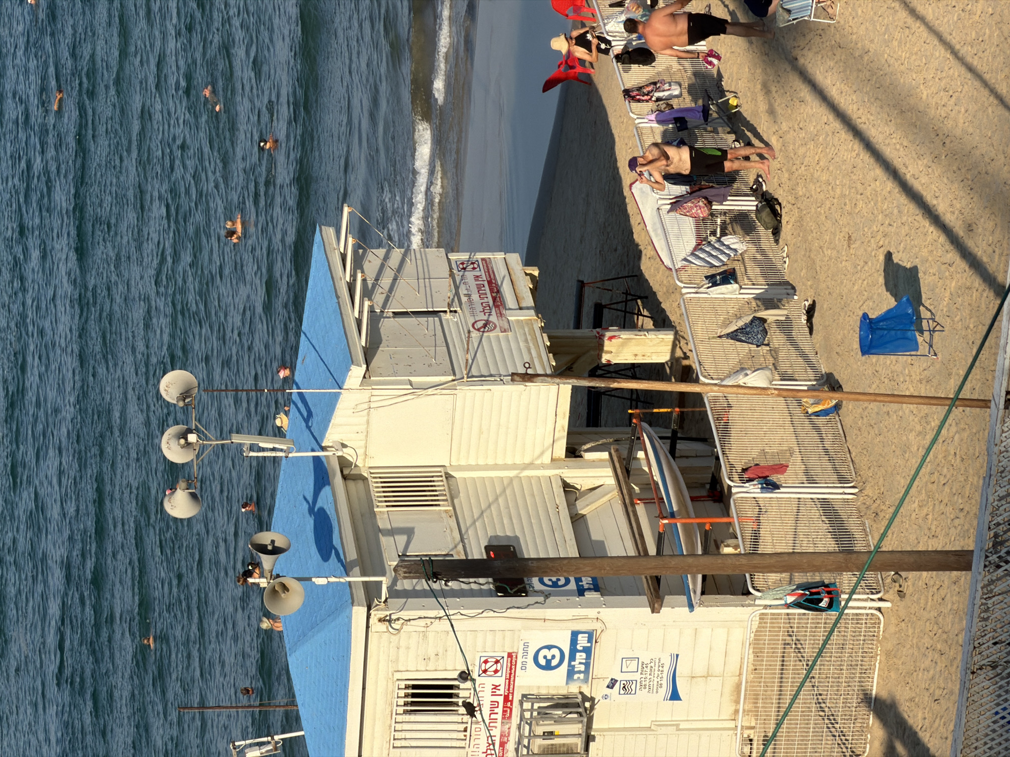 This is a high-angle, medium shot of a beach in Bat Yam, Israel, on a sunny day. The scene is dominated by a white lifeguard station with a blue roof. Several people are visible in the water, swimming or wading. On the sandy beach, there are a few individuals, some wearing swimwear. One man, shirtless and wearing black swim shorts, is standing and looking towards the water. Another man, also shirtless and in black swim shorts, is walking away from the camera. There are several beach chairs, umbrellas, and bags scattered on the sand. A fence runs along the front of the lifeguard station, with towels and clothing hanging on it. A small boat is visible on a rack behind the fence. The ocean is calm with gentle waves. The overall atmosphere is one of a typical summer day at the beach.