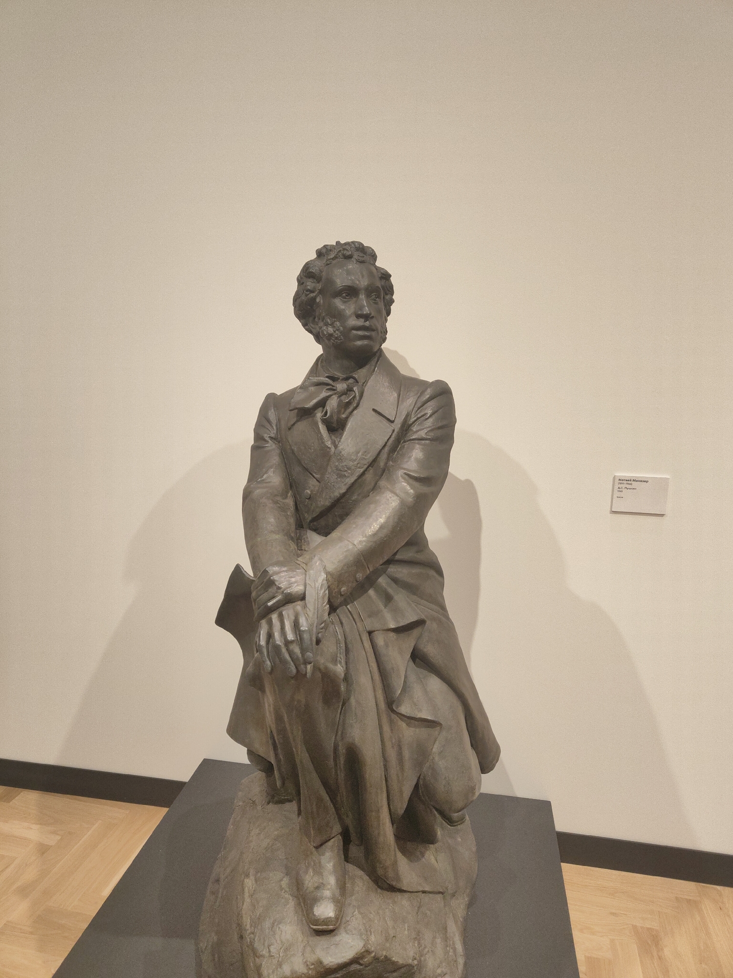 The image shows a bronze statue of a man in a suit. He is seated, with his hands clasped on his knee. The statue is displayed on a gray plinth in a museum setting. A label is affixed to the wall behind the statue. The wall is light beige, and the floor is made of wood. The location is unknown, Unknown.