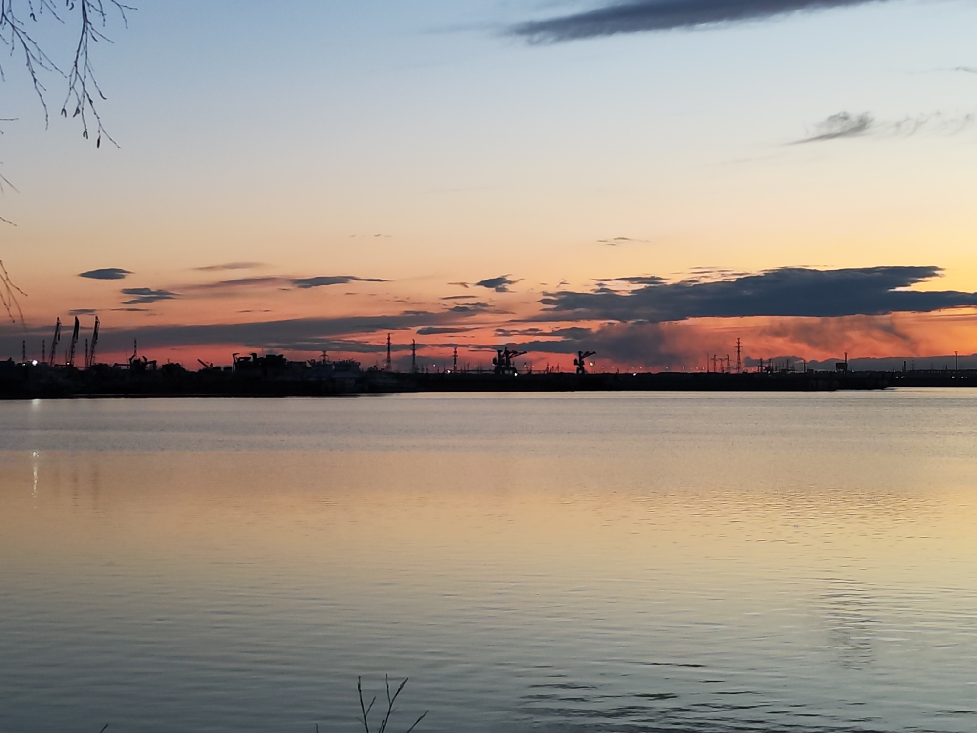 This image presents a tranquil waterfront scene at either sunrise or sunset, set against the backdrop of an industrial horizon in Chaikovsky, Russia. The sky dominates the upper half of the frame, displaying a gradient of colors that transition from a light, clear blue at the top to soft yellows, oranges, and vibrant pinks and purples closer to the horizon. Several elongated, dark clouds are scattered across the sky, particularly concentrated above the distant landmass, with their undersides illuminated by the low sun.
On the distant horizon, a dark, silhouetted industrial or urban area stretches across the frame. This skyline is characterized by numerous structures, including tall cranes, various buildings, and possibly smokestacks, indicating a working port or factory complex situated by the water. There appears to be a faint haze or smoke rising above parts of this industrial zone.
The lower half of the image is occupied by a calm body of water, likely a lake or wide river, which beautifully reflects the warm, colorful hues of the sky and the dark silhouettes of the distant structures. Subtle ripples disturb the water's surface, creating a dynamic yet serene reflection. In the top left corner, the delicate, bare branches of a tree extend into the frame, and a few similar branches are visible at the very bottom, adding a natural foreground element. The overall impression is one of peaceful contemplation of a twilight landscape where nature meets industry. No text is visible in the image.