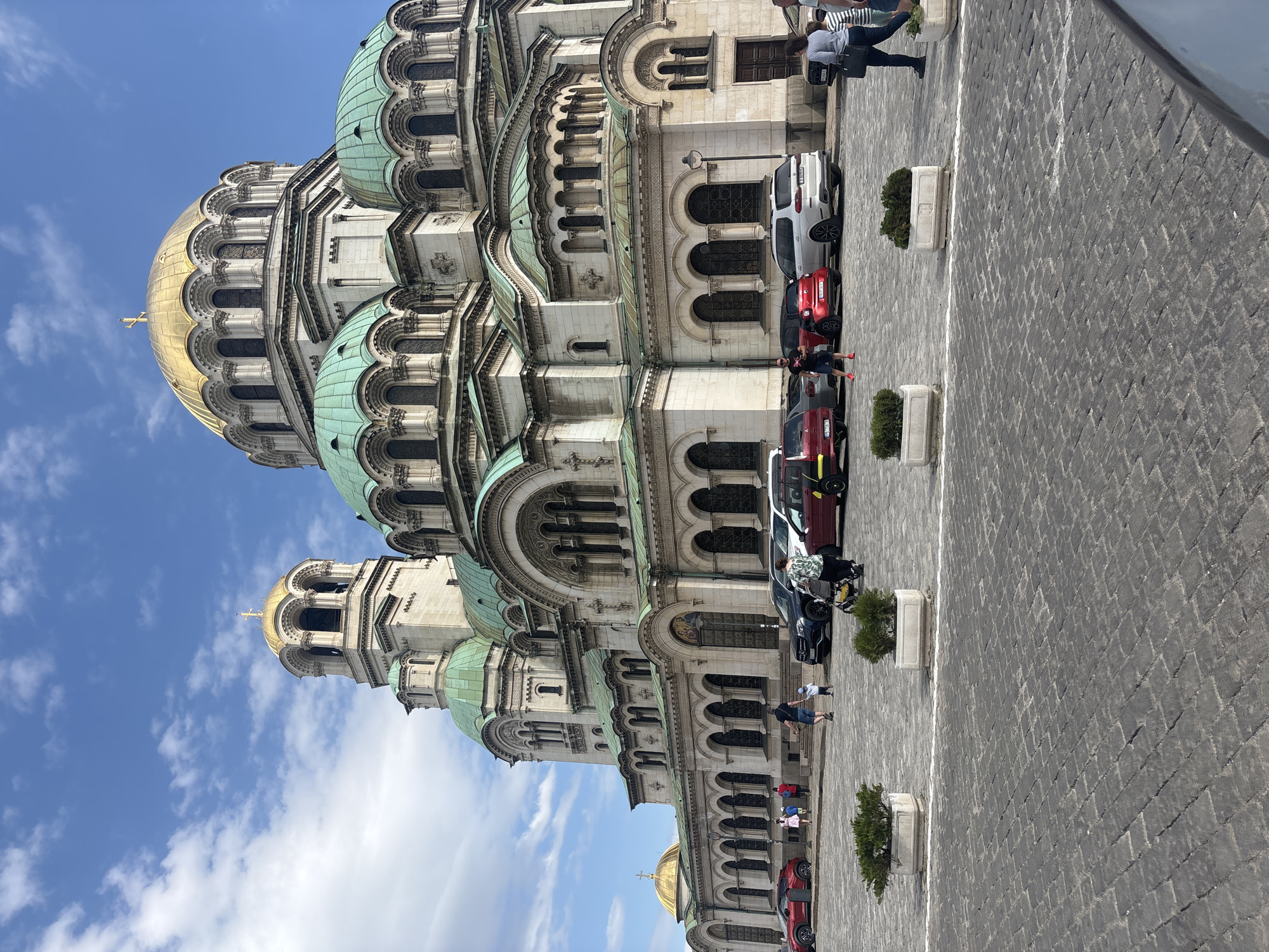 The image shows the Alexander Nevsky Cathedral in Sofia, Bulgaria. The cathedral, with its prominent gold and green domes, stands tall against a bright blue sky with scattered clouds. The façade of the building is ornate and detailed, featuring arches and windows. In front of the cathedral, a paved square is visible. Parked cars are aligned along the building's side. People can be seen walking nearby the cars. Small potted plants are placed intermittently along the square.