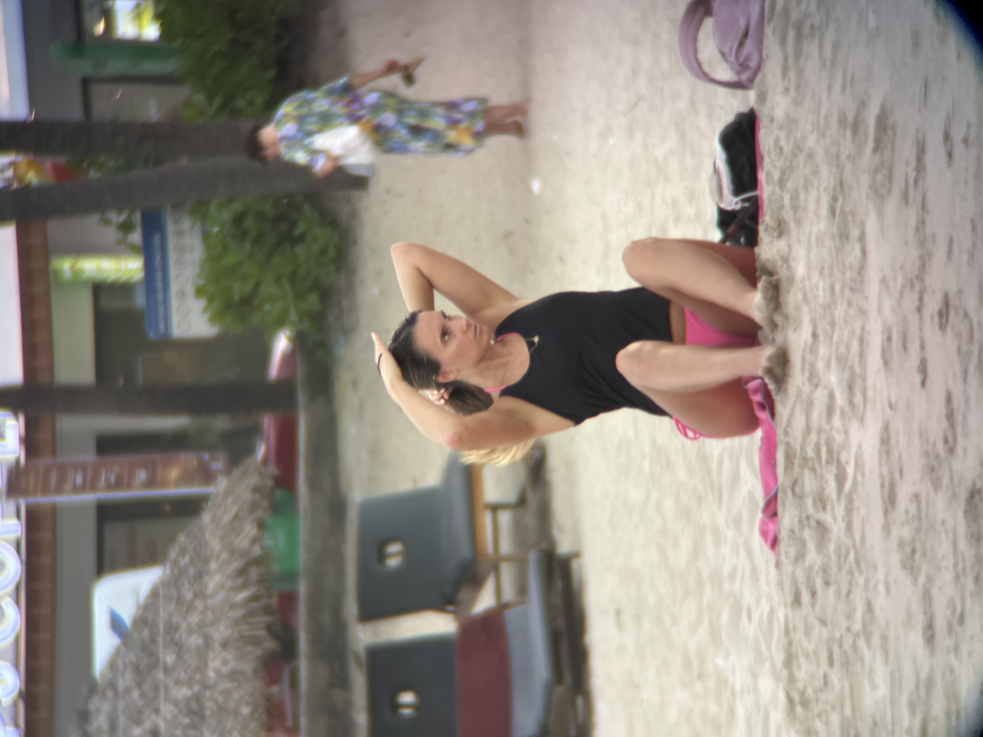 An outdoor, eye-level, medium shot captures a young woman sitting on a sandy beach. She is wearing a black tank top and pink bikini bottoms. Her hair is pulled back, and she is touching her head. Her legs are crossed in front of her, and her feet are visible. There is a pink towel to her left and a purple bag to her right. In the background, two empty lounge chairs are visible, and beyond them, a person in a brightly colored floral dress is walking away from the camera. The scene is set in Đà Nẵng, Vietnam, a coastal city known for its beaches and vibrant atmosphere. The beach appears to be a public area, with elements of commercial activity, such as a thatched-roof umbrella and what appears to be a building with signage, visible in the far background. The lighting suggests it is daytime, possibly late afternoon, with soft, diffused light.