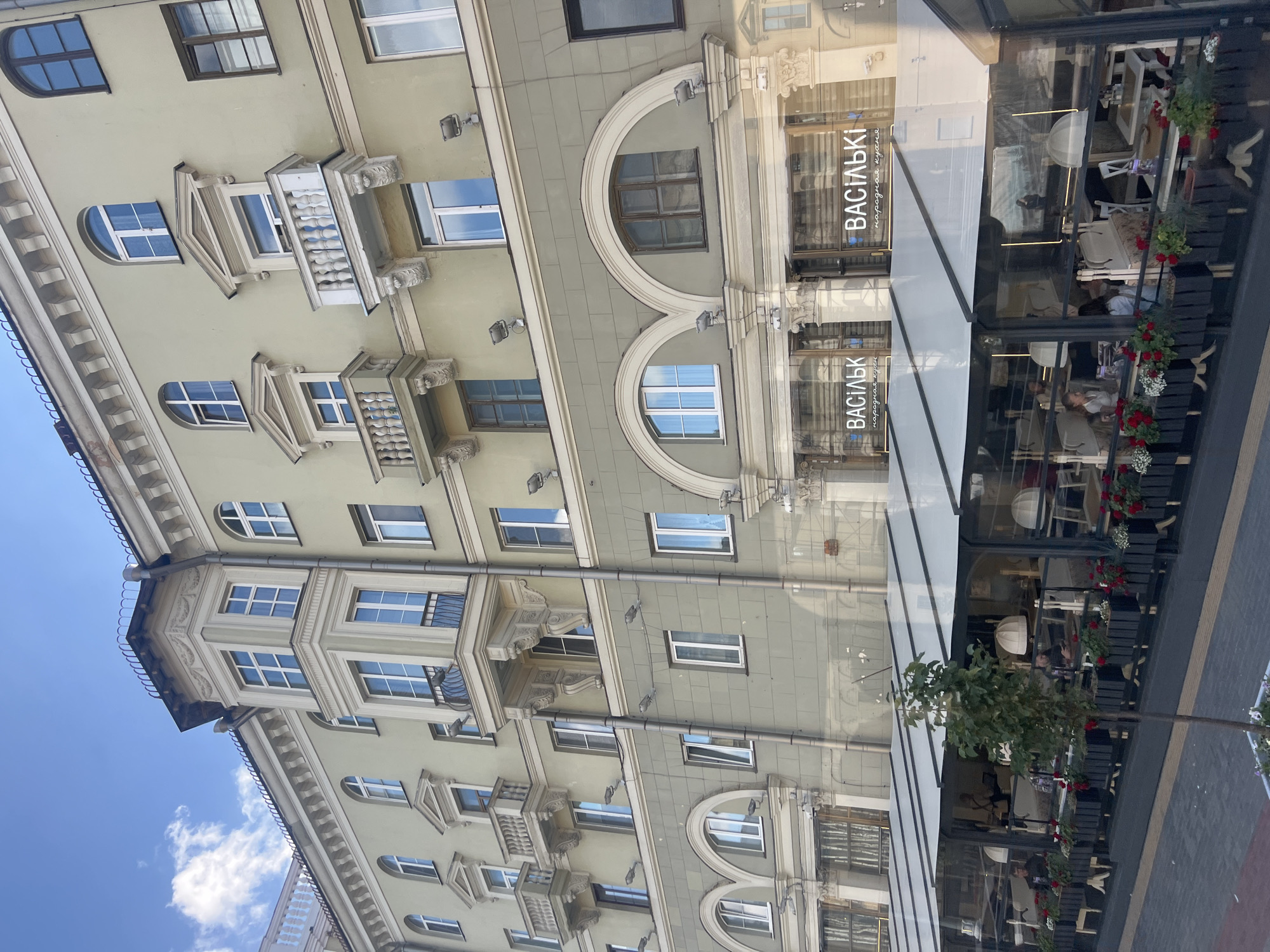 A full-shot, eye-level, exterior photograph captures a pale yellow and muted green multi-story building with a restaurant terrace in front. The architecture exhibits classical details, including arched windows, balconies with ornate railings, and decorative cornices. The building has multiple windows, some with arched tops, and small balconies with wrought-iron railings are visible on the upper floors. The ground floor features a modern, glass-enclosed restaurant terrace with tables and chairs visible inside. Two signs on the facade read "ВАСІЛЬКІ" and "народная кухня" (Vasilki and National Cuisine). The terrace is adorned with potted plants and red flowers in planters. A young, slender tree stands in a planter in front of the terrace. The sky is blue with scattered white clouds. The scene appears to be a daytime shot in an urban setting, likely a street in Minsk, Belarus, given the Cyrillic signage.