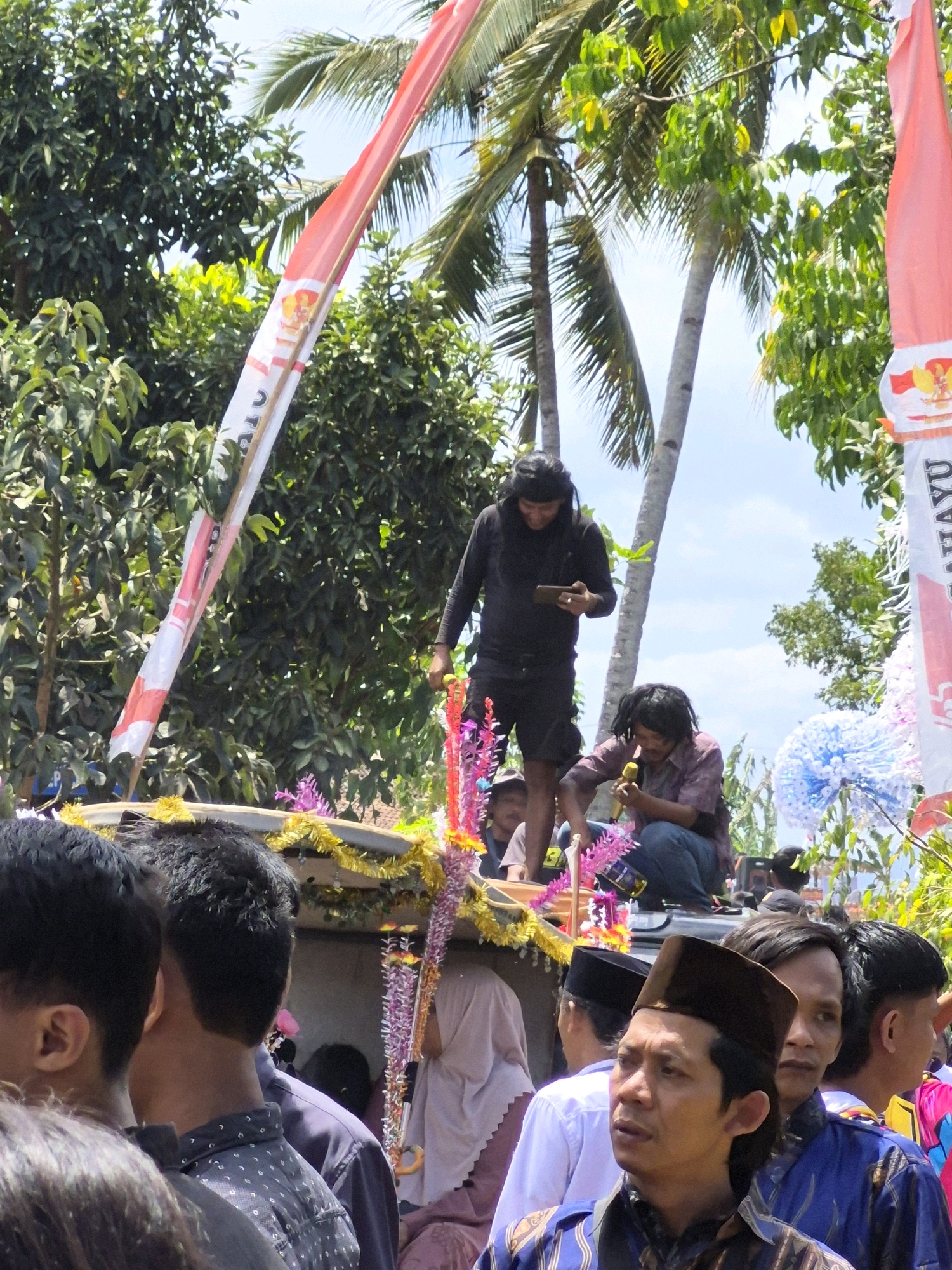 The image depicts an outdoor gathering, likely a cultural event or celebration, in Grantung, Indonesia. The foreground is filled with the backs of several men, suggesting they are part of a crowd watching something. One man in the mid-ground, wearing a brown cap, is looking to his left with a neutral expression. Further back, a person on a raised platform is wearing a black shirt and shorts and holding a phone. Another person next to them is kneeling and holding a yellow object, possibly a microphone or a musical instrument. Decorative banners, one red and white and another with red and white stripes and crests, are prominently displayed, suggesting a national or organizational affiliation. Palm trees and lush green foliage frame the scene, indicating a tropical or subtropical environment. The presence of colorful decorations on the platform suggests a festive atmosphere.