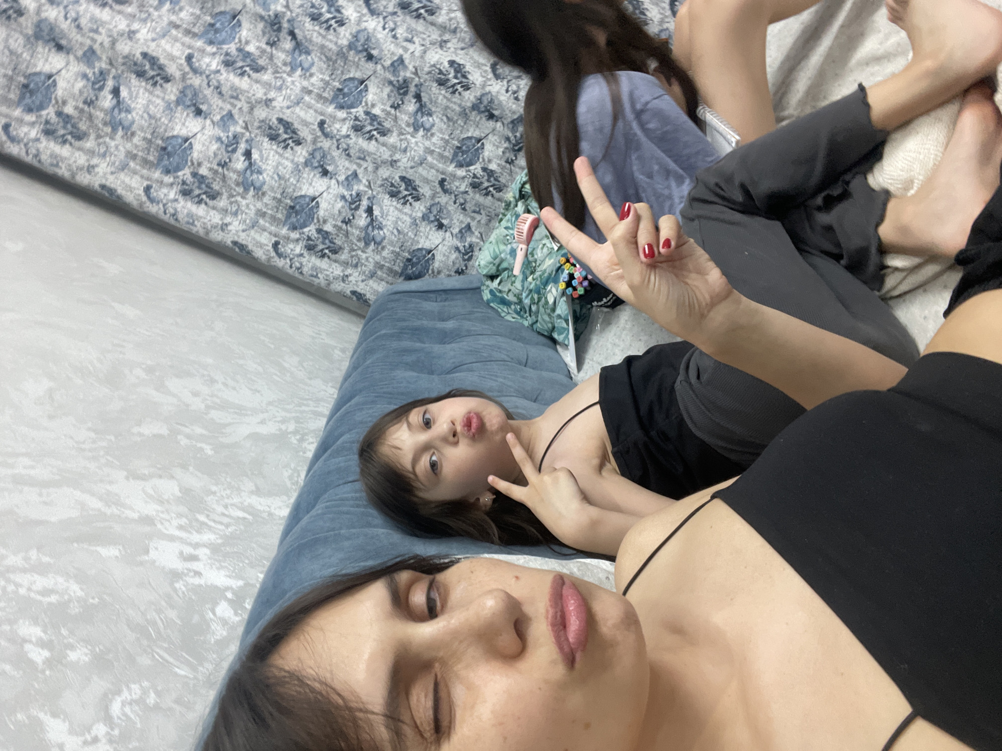 A selfie taken from a low angle shows a woman and a child, presumably female, lying on a blue futon. A second individual is partially visible behind them. The woman in the foreground is fair-skinned and wearing a black tank top. The child is lying behind her and to the left, also making a peace sign. The child is wearing a black tank top as well. In the background, a third person, potentially an older child or teenager, is partially visible near the head of the futon. The futon is in a room with white walls and light-colored flooring. A bed with a blue and white patterned comforter is in the background. The location is presumed to be in Izhevsk, Russia.