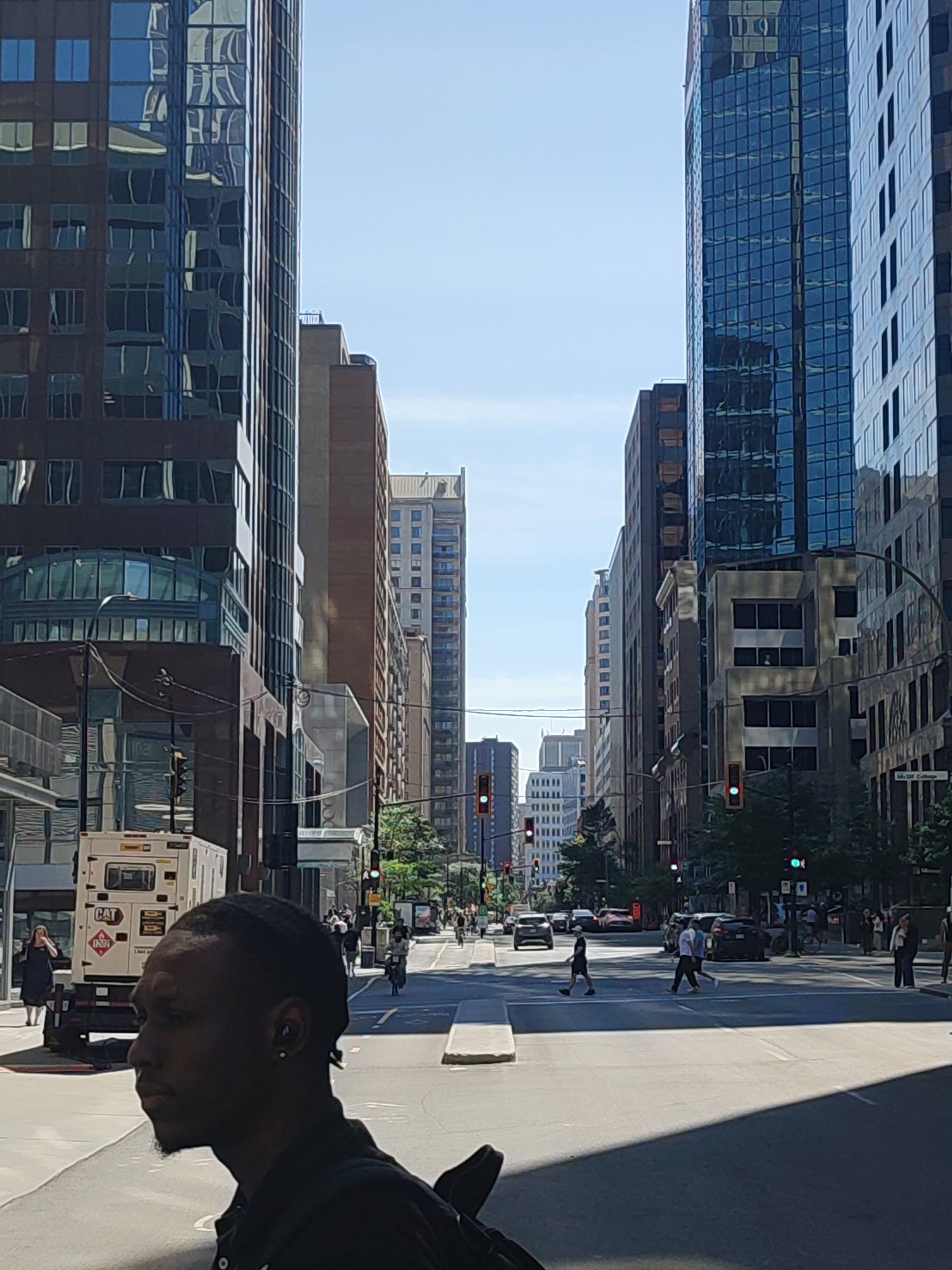 The image captures a street scene in Montreal, Canada, under a clear blue sky on a sunny day. The foreground is dominated by the silhouetted profile of a Black man facing left, wearing a dark shirt and an earring. Behind him, a white generator truck with "CAT" branding is parked on the left side of the street. The street itself is paved, with crosswalks indicating pedestrian crossings.
The background features a canyon of tall, modern glass and concrete buildings characteristic of a downtown urban environment. The architecture is varied, with some buildings displaying dark reflective glass facades and others with lighter-colored materials. Traffic lights are visible at intersections, with some showing green lights. Pedestrians are seen crossing the street at designated areas, and vehicles, including cars and a bicycle, are present on the road. Trees line parts of the sidewalk, adding touches of green to the otherwise predominantly grey and blue cityscape. The perspective suggests the photographer is standing on a street corner or sidewalk, looking down the length of the street.