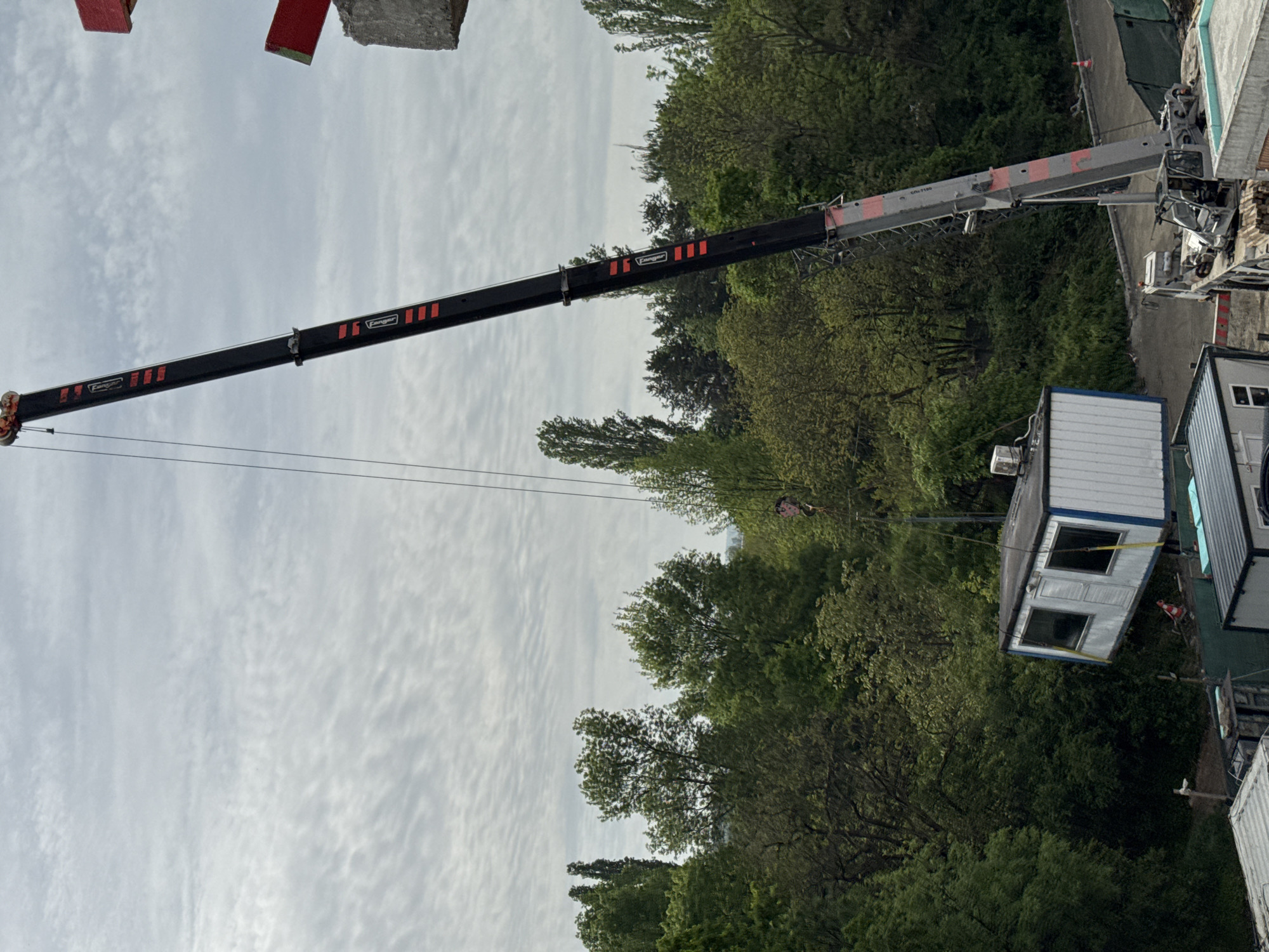 A crane is lifting a white portable building in Unknown, Unknown. The crane, with a black boom and red markings, extends from the top right of the frame towards the left, its lifting cable attached to the building. The portable building has windows and blue trim. The background consists of trees and a cloudy sky.
