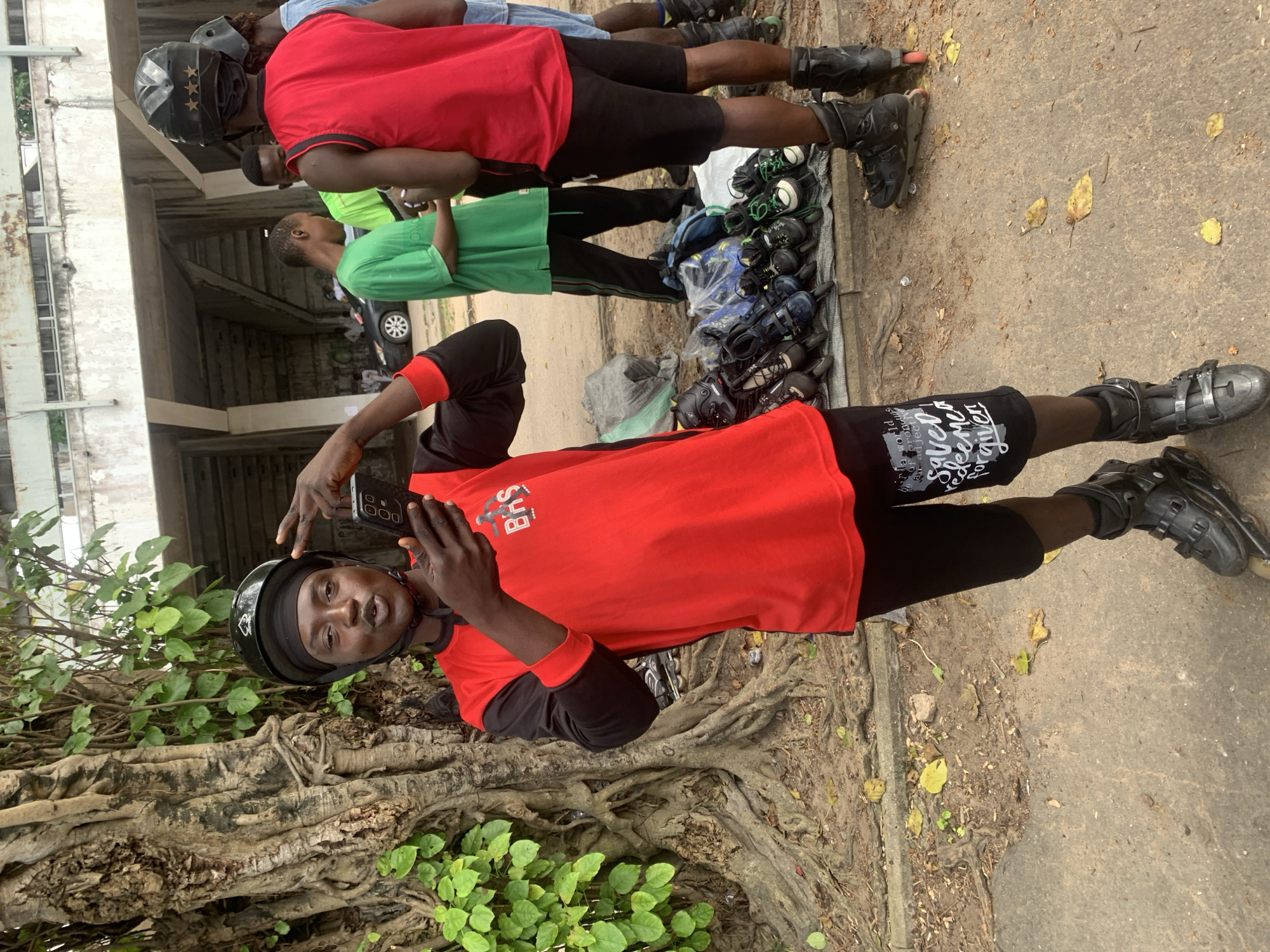 In Alagbede, Nigeria, a young Black man wearing a red long-sleeved shirt with black sleeves, black shorts with white lettering, a black helmet, and black rollerblades poses for a picture. He holds a black smartphone in his right hand and makes a peace sign with his left hand, looking towards the camera with a playful expression. Behind him, a large tree with exposed roots is visible to his left, and to his right, a row of rollerblades is displayed on the ground, suggesting a marketplace or rental service. Several other individuals, also wearing casual attire and some with helmets, are present in the background, with a concrete structure resembling bleachers or a covered walkway in the distance. The ground is a mixture of dirt and concrete, with scattered leaves and debris. The scene appears to be outdoors, under daylight conditions.
