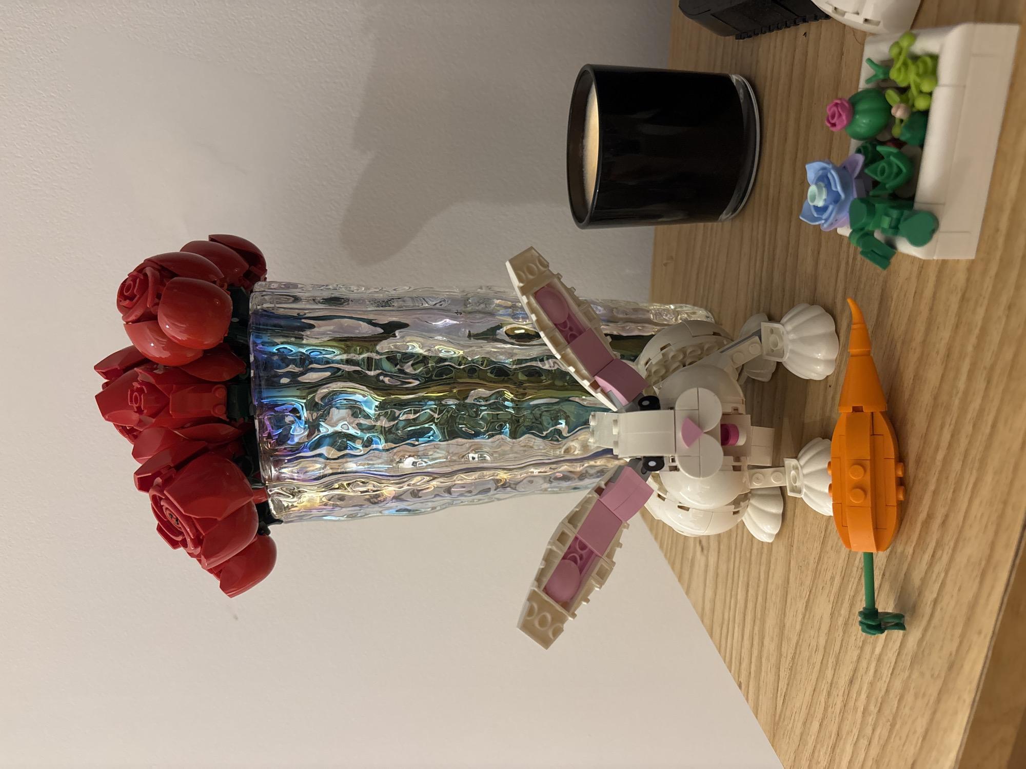 The image depicts a still life arrangement. Dominating the scene is a vase filled with a bouquet of red roses. Adjacent to the vase is a Lego rabbit figurine holding a carrot. Behind the rabbit is a black candle and a Lego flowerpot with blue and pink flowers. The arrangement rests on a wooden surface. The setting does not appear to be in Buenos Aires.