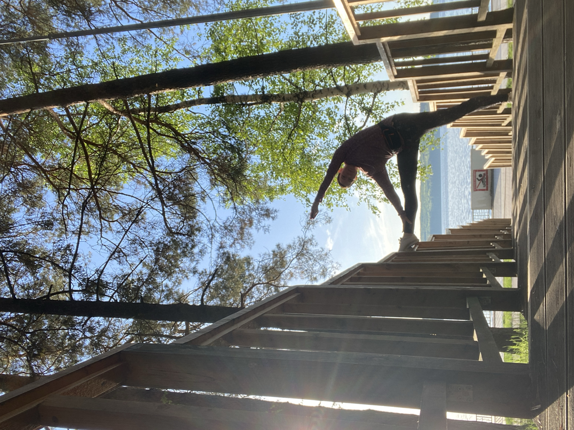 A person is seen performing yoga on a set of wooden stairs next to a body of water. The location is Chaikovsky, Russia, near the Kama River. The wooden stairs are part of a public infrastructure, like a boardwalk or viewing platform, bordered by a wooden railing. The trees in the background suggest a natural environment. The person is in the process of doing the triangle pose.