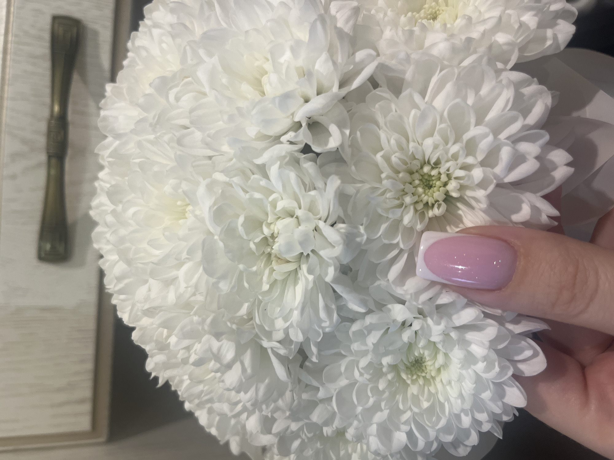 An extreme close-up shot focuses on a bouquet of white chrysanthemum flowers. A person's hand, with a light pink French manicure, is gently touching one of the flowers. In the background, a light-colored wooden cabinet with a brass handle is visible. The setting appears to be indoors, possibly a home or a florist shop. There is no specific information to suggest this scene is located in Tyumen, Russia.