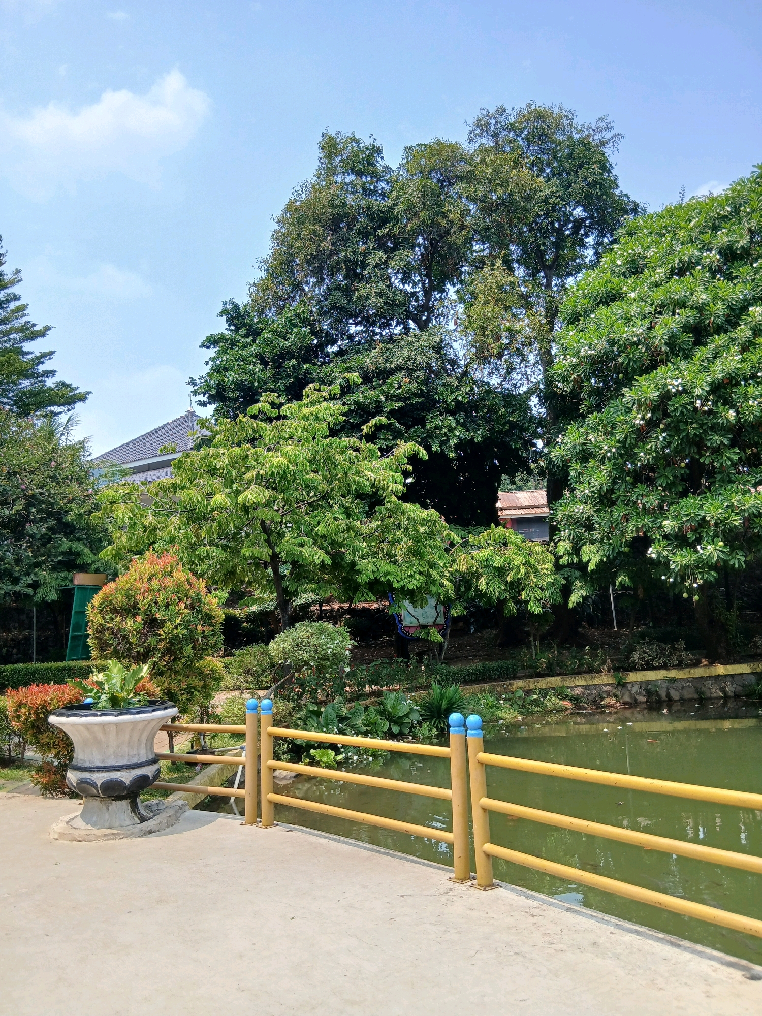This is a photograph of a scenic area in Depok, Indonesia, likely a public park or garden, showcasing a tranquil waterscape surrounded by lush greenery under a clear blue sky with scattered clouds. In the foreground, a concrete pathway leads the eye towards a vibrant yellow railing with blue-capped posts, bordering a calm, green body of water. To the left of the pathway, a large, ornate stone planter holds a variety of plants, adding a touch of decorative horticulture. The background is dominated by a dense collection of trees of various sizes and species, creating a verdant backdrop. Some of these trees have abundant foliage, while others, particularly a large tree on the right, are adorned with small white flowers, suggesting a blooming season. A glimpse of a building with a dark, tiled roof can be seen peeking through the trees on the left, hinting at the presence of man-made structures within this natural setting. The overall impression is one of a peaceful and well-maintained outdoor space, inviting relaxation and enjoyment of nature.