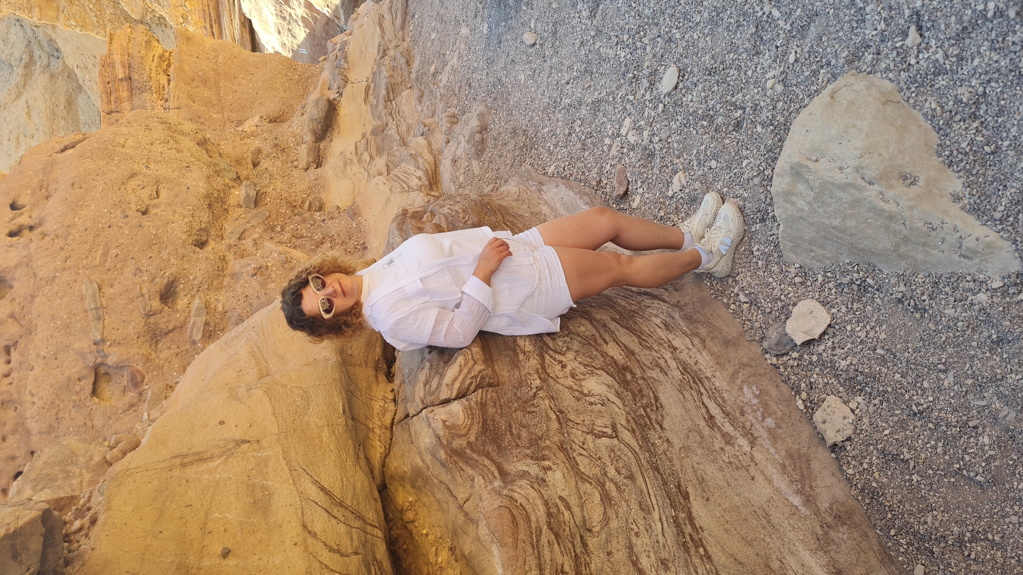 A woman is lying on a patterned rock in a canyon. She is wearing a white shirt, white shorts, white socks and off-white tennis shoes, and sunglasses. The surrounding environment includes canyon walls on the left and rocky terrain on the right. The location is in Hevel Eilot Regional Council, Israel.