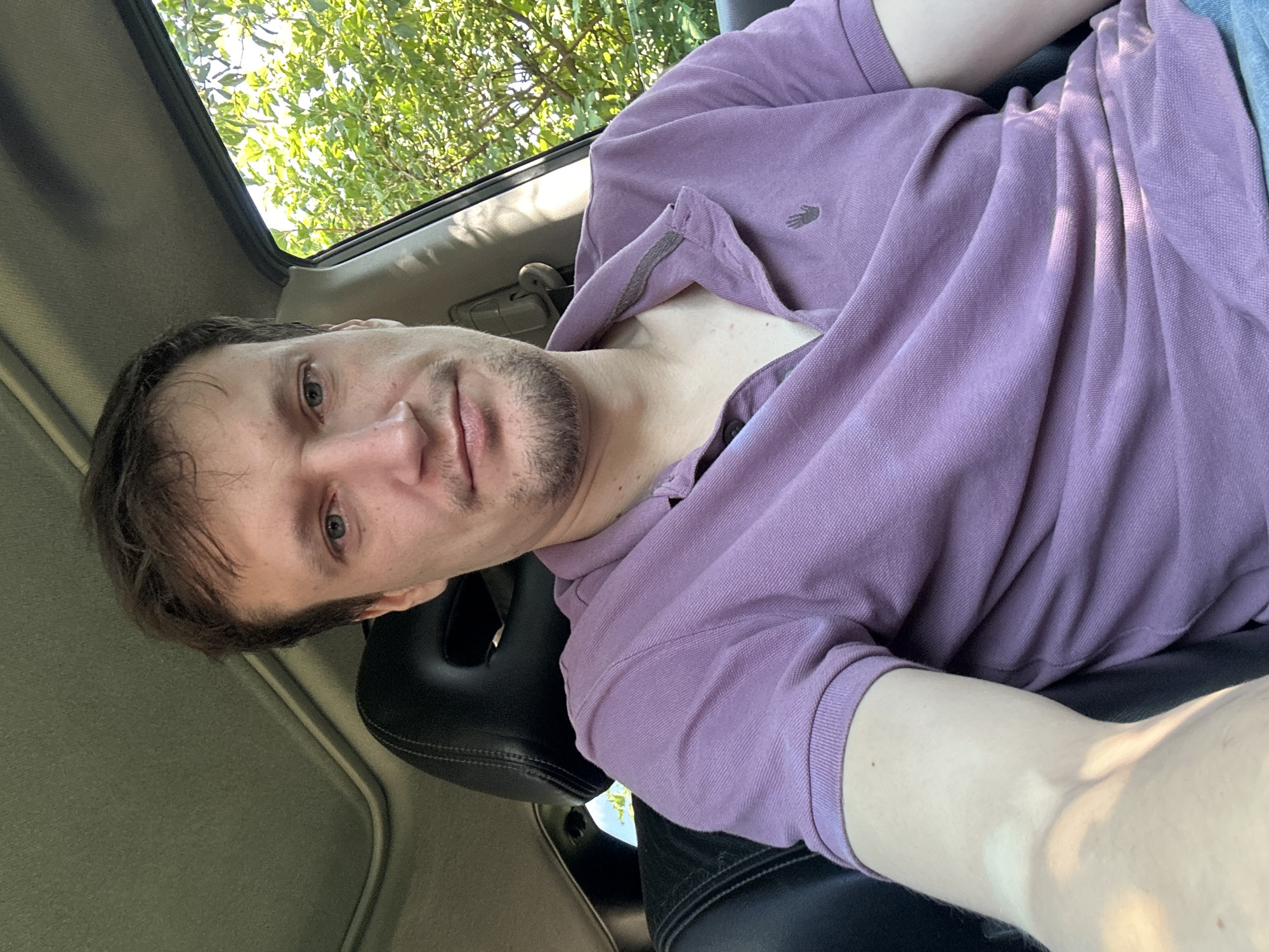 A man sits in the driver's seat of a car, looking directly at the camera. He has light skin, brown hair, and a light beard. He is wearing a purple polo shirt with a small black emblem on the left side of the chest. The car's interior is visible, including a black leather headrest and a sunroof. Outside the car, green trees and foliage are visible, suggesting a daytime setting, possibly in a rural or suburban area. The location is Shatava, Ukraine.