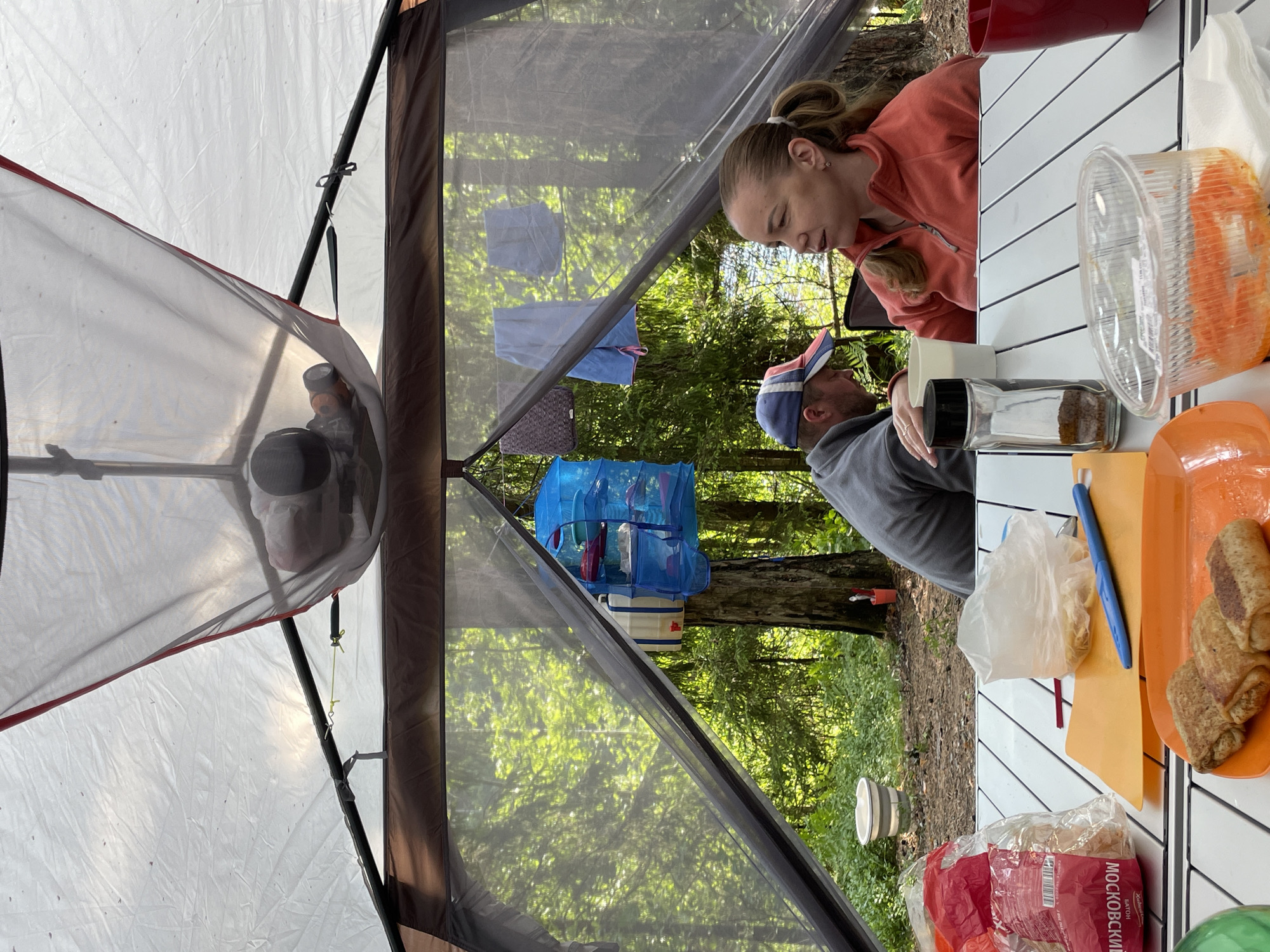 In Roshchino, Russia, a scene unfolds within a large tent, likely at a campsite given the surrounding forest and natural light. Two adults are present at a white folding table. A man wearing a red, white, and blue baseball cap is seated, with his left hand resting on the table. A woman, with blonde hair pulled back in a ponytail, is sitting opposite him, wearing an orange fleece jacket and looking to her left. On the table, there are various items including a bag of food with Russian text, a clear plastic container with food, a spice grinder, a glass of water, and an orange plate with three rolled food items. In the background, through the mesh of the tent, a lush green forest with tall trees is visible. There is also a blue hanging organizer with items inside, a water jug, and a white bucket. Above the people, a netted hammock is suspended, containing what appears to be a sleeping child. The overall atmosphere suggests a casual outdoor gathering or camping trip.