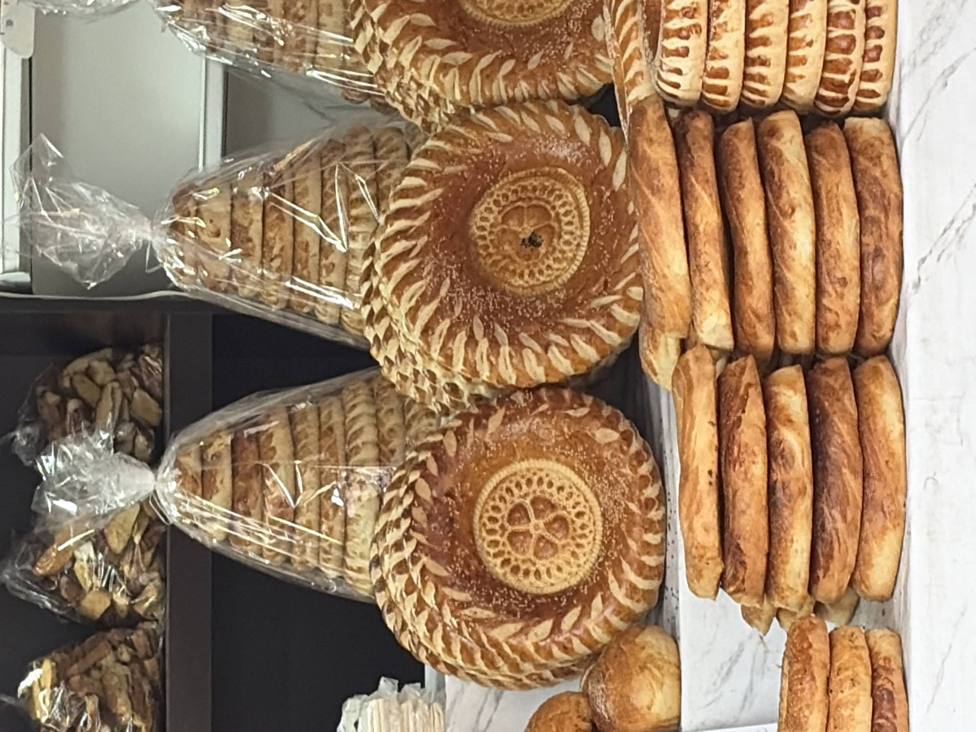 A counter displays a variety of baked goods in what appears to be a bakery. In the foreground, to the left, are stacks of flatbreads and some smaller, round loaves. To the right, there are stacks of flatbreads and also some larger, round, decorative breads. Behind these, there are two tall, conical stacks of bread wrapped in clear plastic. Further back, on a shelf, are bags of bread-like items. The overall impression is of a well-stocked bakery offering a range of bread products.