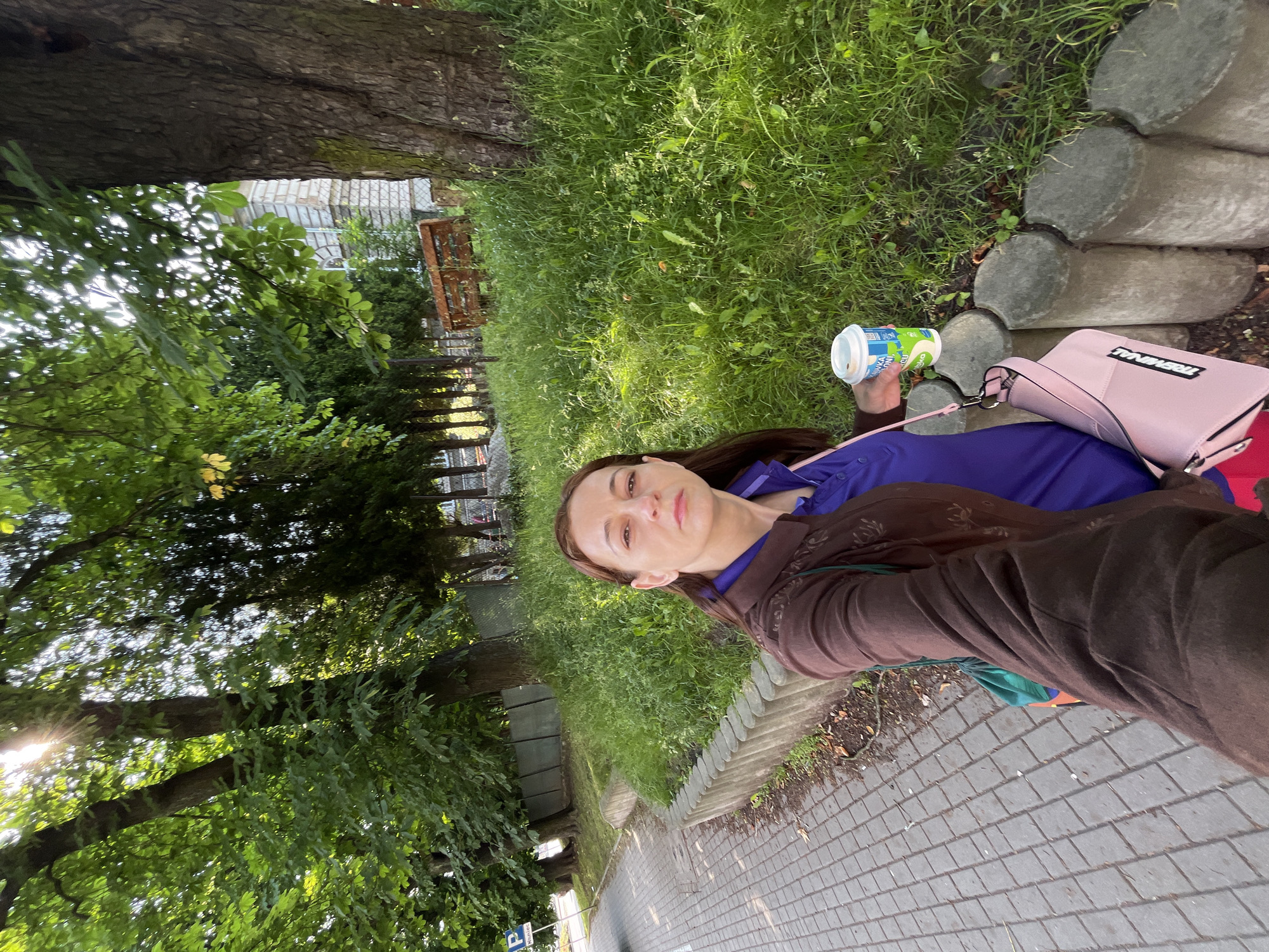 A woman is standing on a sidewalk next to a grassy area with trees. She is wearing a brown jacket over a purple shirt and is holding a cup of yogurt. A pink purse is slung over her shoulder. There is a wooden bench and a building in the background. The sun is shining through the trees.